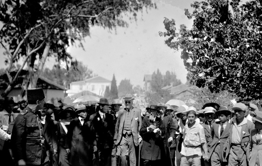 در این تصویر، گروهی از مردم در یک فضای باز دیده می‌شوند. بخشی از آنها چترهایی در دست دارند و ظاهراً در حال تماشای یک رویداد یا شخصیت مهم هستند. در وسط تصویر، مردی با عصا و لباس رسمی در حال حرکت است. اطراف او جمعیتی از مردان و زنان ایستاده‌اند که همگی لباس‌های رسمی و مرتب به تن دارند. در سمت چپ، فردی در لباس نظامی دیده می‌شود که به نظر می‌رسد به عنوان نگهبان یا محافظ عمل می‌کند. فضای عمومی و هوای آفتابی نشان‌دهنده یک روز شلوغ و پرحادثه است.