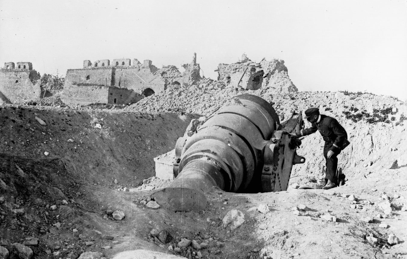 L'image montre un homme travaillant près d'un grand canon ou d'une pièce d'artillerie en position, probablement dans un environnement de guerre. À l'arrière-plan, on peut voir des bâtiments en ruine, ce qui suggère que la scène se déroule dans une zone de conflit, possiblement après un bombardement. Le paysage est aride et rempli de débris, ce qui accentue l'atmosphère de destruction. L'homme semble concentré sur son travail, illustrant les efforts militaires dans un contexte difficile.