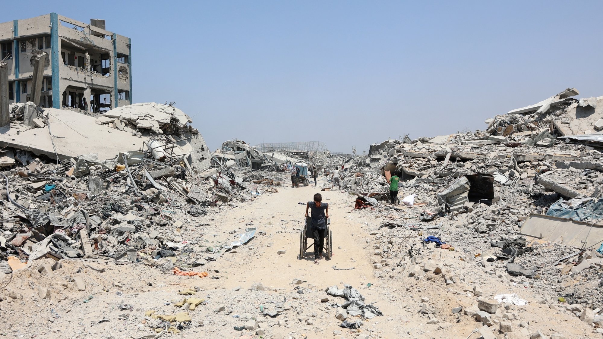 A person in a wheelchair navigates a debris-filled street amid collapsed buildings.