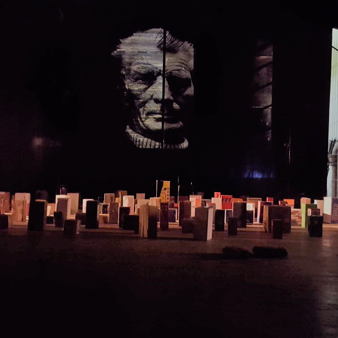 L'image montre une scène intérieure avec un grand mur sombre sur lequel se trouve un portrait en noir et blanc d'une personne âgée. Au premier plan, il y a un sol rempli de petites structures ressemblant à des livres ou des modules disposés de manière organisée. À l'arrière-plan, on peut apercevoir une personne assise, probablement en train de travailler ou de se préparer. L'éclairage est faible, créant une atmosphère mystérieuse.