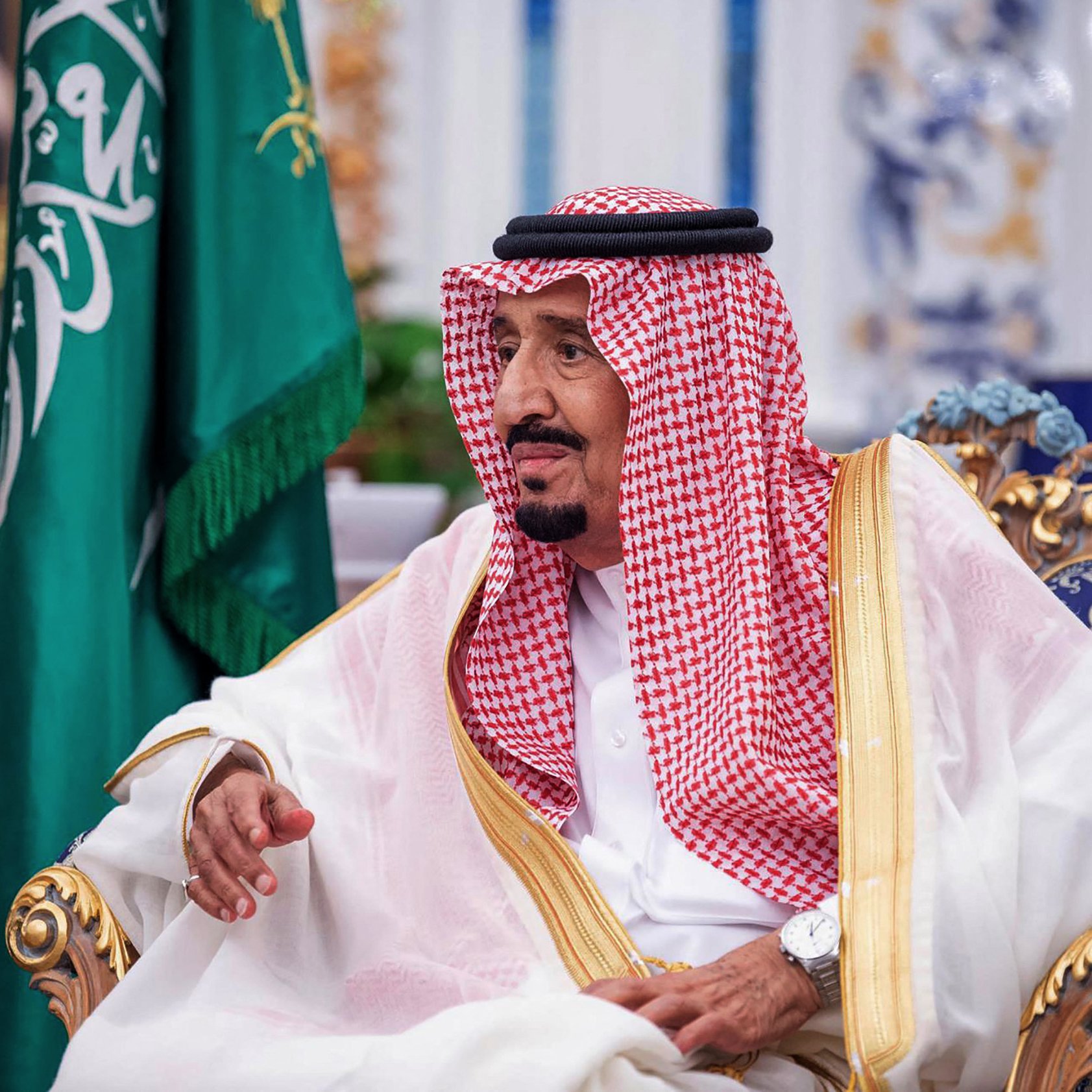 L'image montre un homme assis sur un fauteuil orné, vêtu d'une robe traditionnelle appelée "thobe" et couvert d'un "ghutrah", un foulard. Il est entouré d'une décoration élégante avec des éléments représentant la culture arabe. En arrière-plan, on peut voir un drapeau saoudien, ce qui suggère qu'il pourrait s'agir d'une figure importante ou d'un leader. L'expression de son visage indique qu'il est en plein échange ou réflexion.