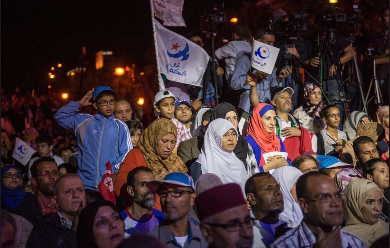 L'image montre une foule rassemblée lors d'un événement, probablement politique. On peut voir des personnes de différents âges, y compris des enfants. Certaines personnes portent des drapeaux avec des symboles, suggérant qu'elles soutiennent un mouvement ou un parti. L'atmosphère semble intense, avec des caméras et des journalistes présents, indiquant l'importance de l'événement. Les gens affichent diverses expressions, allant de la concentration à l'enthousiasme.