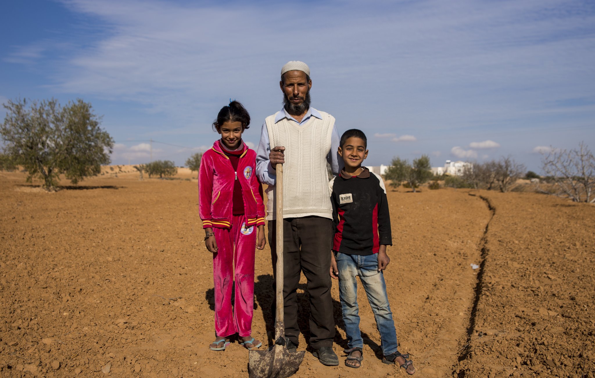 L'image montre un homme debout au milieu d'un paysage désertique, avec deux enfants près de lui, une fille et un garçon. L'homme tient une pelle et porte des vêtements simples. La fille est vêtue d'un ensemble rouge, tandis que le garçon porte un sweat-shirt noir avec des détails rouges. En arrière-plan, on peut voir quelques arbres éparpillés et un ciel bleu clair. Le sol est sec et nu, typique d'une région aride. L'ambiance semble conviviale, suggérant une journée de travail ou de récolte en famille.
