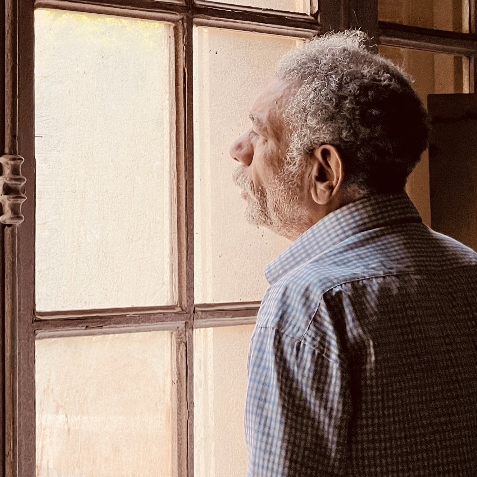 L'image montre un homme âgé qui semble contempler à travers une fenêtre. Il est habillé d'une chemise à motifs et a une posture pensive. La lumière naturalisée entre par la fenêtre, illuminant doucement son visage et créant une ambiance calme. On peut aussi apercevoir des murs en arrière-plan, ajoutant à l'atmosphère de réflexion du moment.