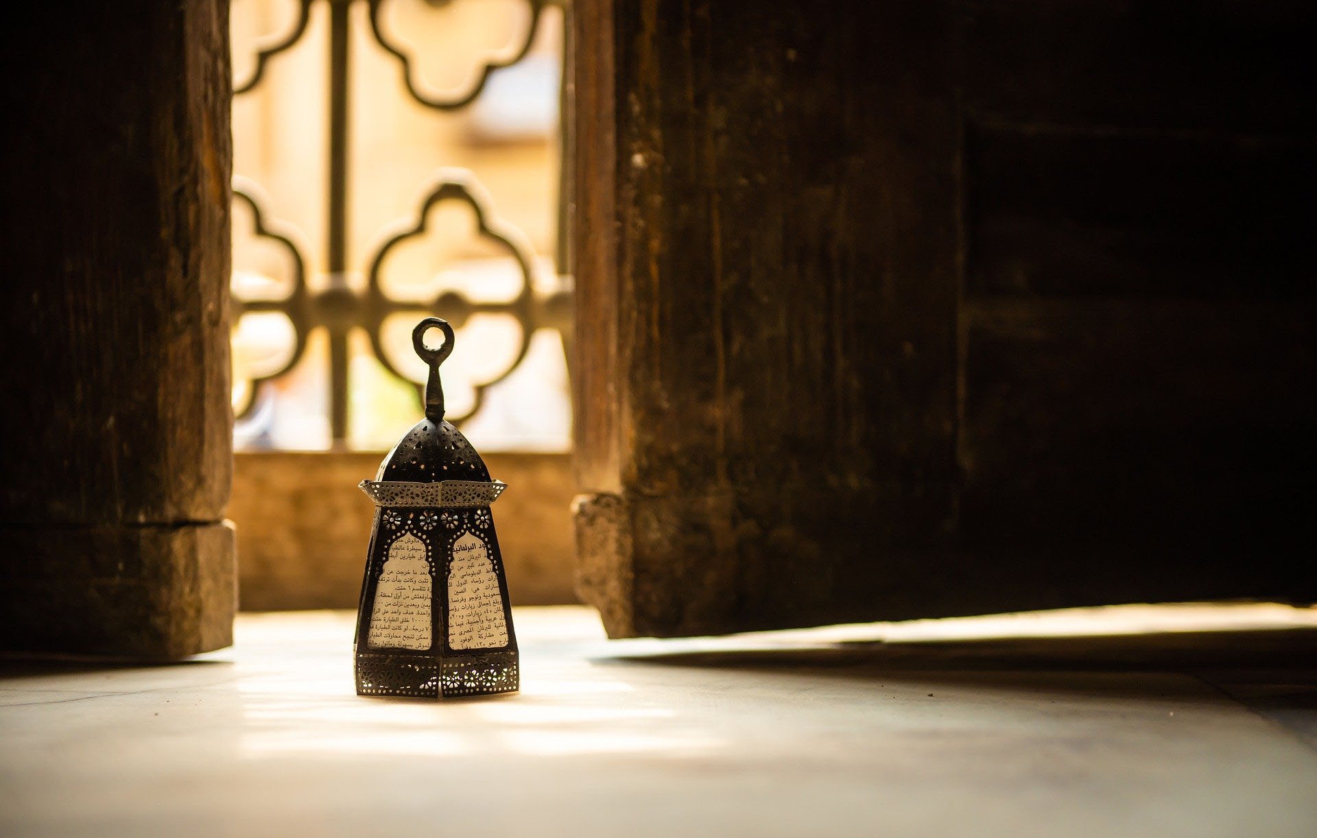 L'image montre une délicate lanterne orientale placée sur le sol, éclairée par une lumière douce, créant une atmosphère chaleureuse. En arrière-plan, on aperçoit un mur avec des motifs décoratifs, et une fenêtre aux formes ornées, laissant transparaître des lumière et ombres intéressantes. La combinaison de la lanterne et de l'environnement évoque une ambiance paisible et méditative.