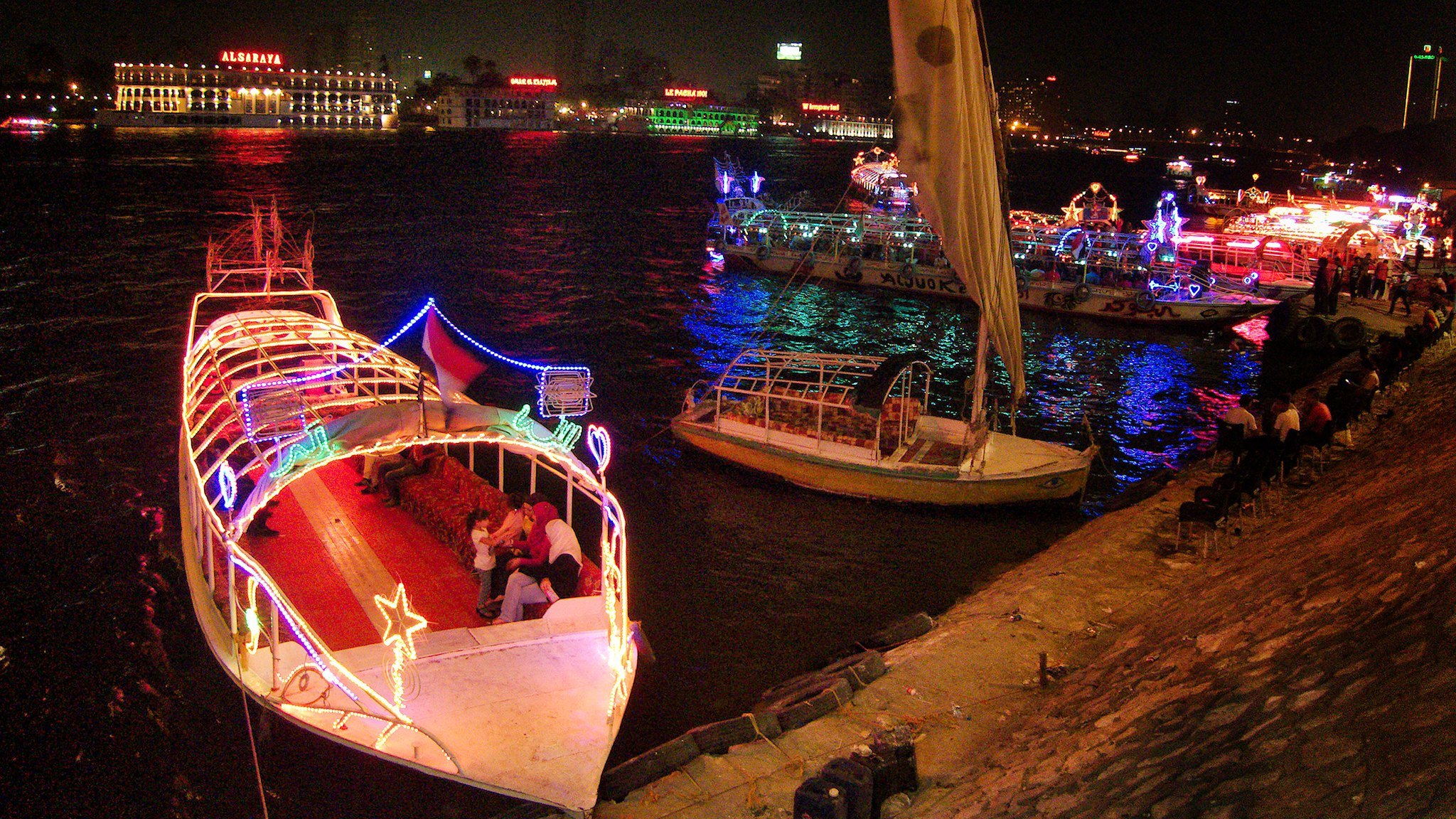 L'image montre un paysage nocturne animé le long d'une rivière. Plusieurs bateaux décorés de lumières colorées sont amarrés, créant une atmosphère festive. On peut voir des gens sur les bateaux et le long de la promenade, profitant de l'ambiance. En arrière-plan, des bâtiments illuminés ajoutent à la scène, avec des reflets dans l'eau. L'ensemble évoque une belle soirée en bord de rivière.