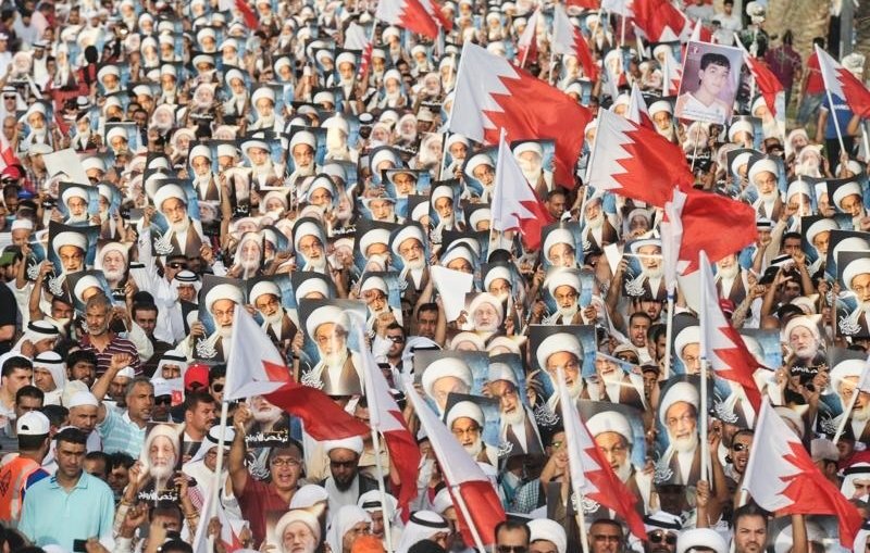 L'image montre une grande foule de manifestants réunis, brandissant des drapeaux et des portraits. Les personnes sont vêtues de manière variée, certaines portant des keffiehs. Les drapeaux représentent probablement le pays en question, tandis que les portraits semblent être ceux de figures clés ou de leaders. L'atmosphère dégage une forte impression de solidarité et de mobilisation autour d'une cause politique ou sociale.
