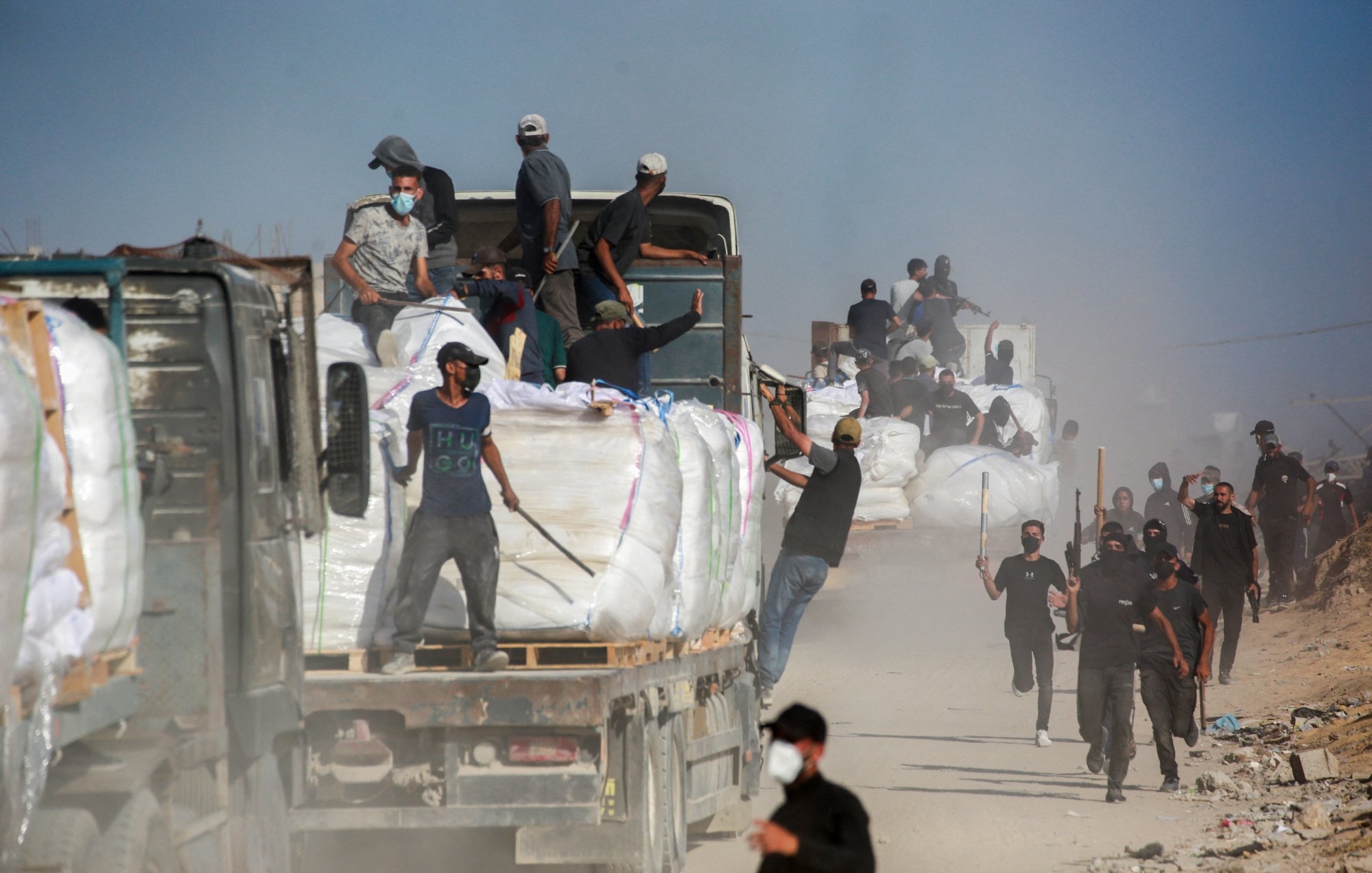تصویر شامل کامیون‌هایی است که بار سنگینی حمل می‌کنند و عده‌ای در حال کار در یک محیط غبارآلود هستند.