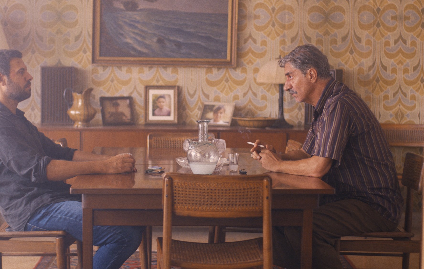L'image montre deux hommes assis à une table en bois dans une pièce décorée avec un papier peint aux motifs rétro. L'ambiance est intérieure et intimiste, avec une lampe à côté et des photos accrochées au mur. Les deux hommes semblent engagés dans une conversation sérieuse. Le premier homme, à gauche, est vêtu d'une chemise sombre et regarde l'autre. Le deuxième homme, à droite, fume une cigarette et semble pensif. L'éclairage doux ajoute à l'atmosphère.
