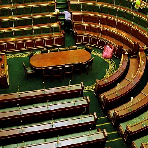 L'image montre l'intérieur d'une assemblée, probablement un parlement ou une salle de réunion officielle. On peut voir des rangées de sièges en bois oblongs disposés en arc de cercle, avec un grand bureau central ovale. Les bancs sont vides, ce qui donne une impression de calme. La décoration est sobre, avec des éléments en bois et un tapis vert. Deux personnes sont visibles, probablement en train de ranger ou d'entretenir l'espace. L'ambiance générale semble formelle et institutionnelle.