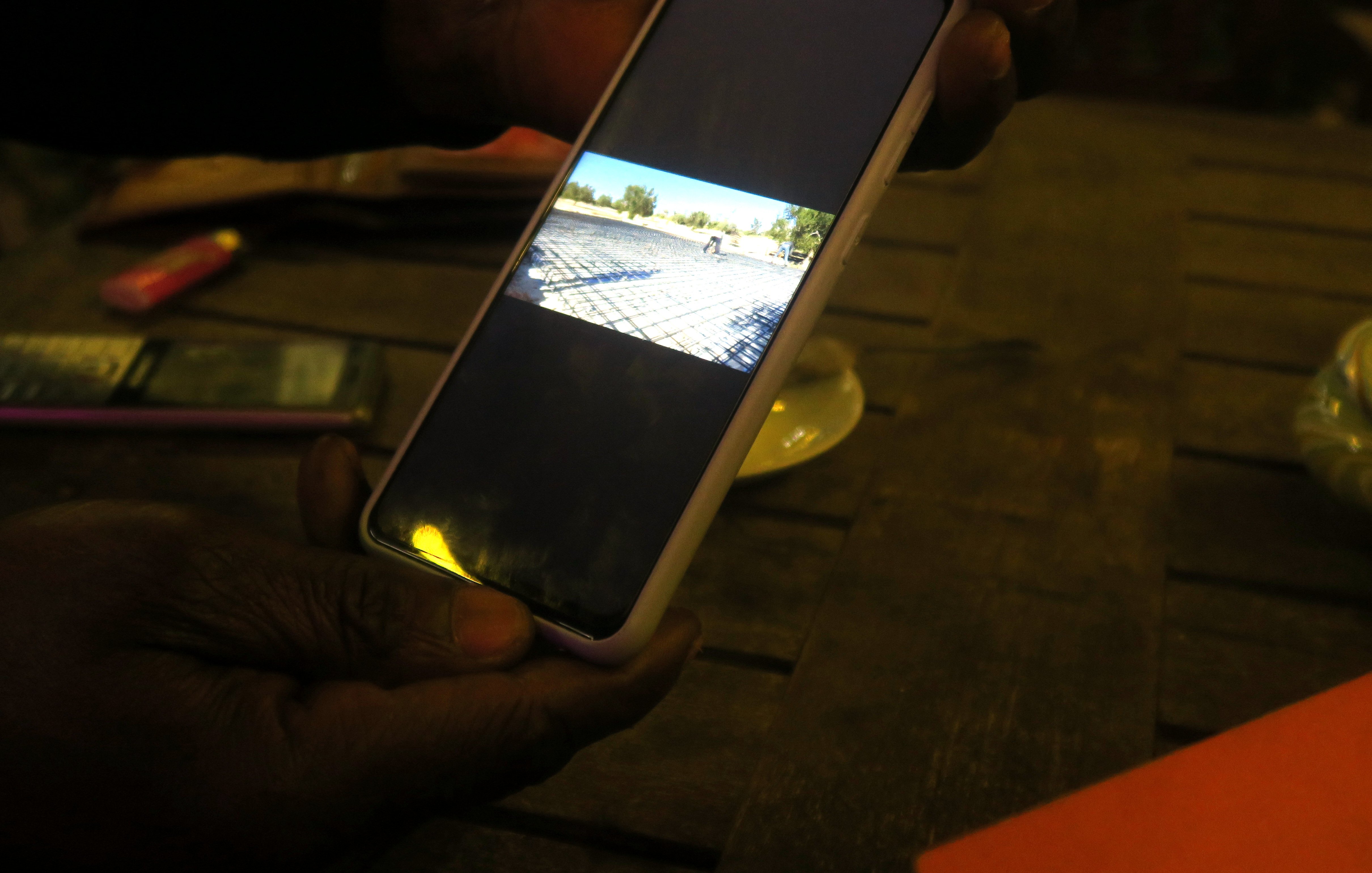 L'image montre une main tenant un téléphone mobile qui affiche une photo sur son écran. La photo visible montre un paysage avec un chemin de pavés et un véhicule au loin. L'éclairage est faible, ce qui donne une ambiance douce à la scène. Le téléphone est positionné sur une table en bois, et l'arrière-plan contient des objets flous, suggérant un environnement convivial.