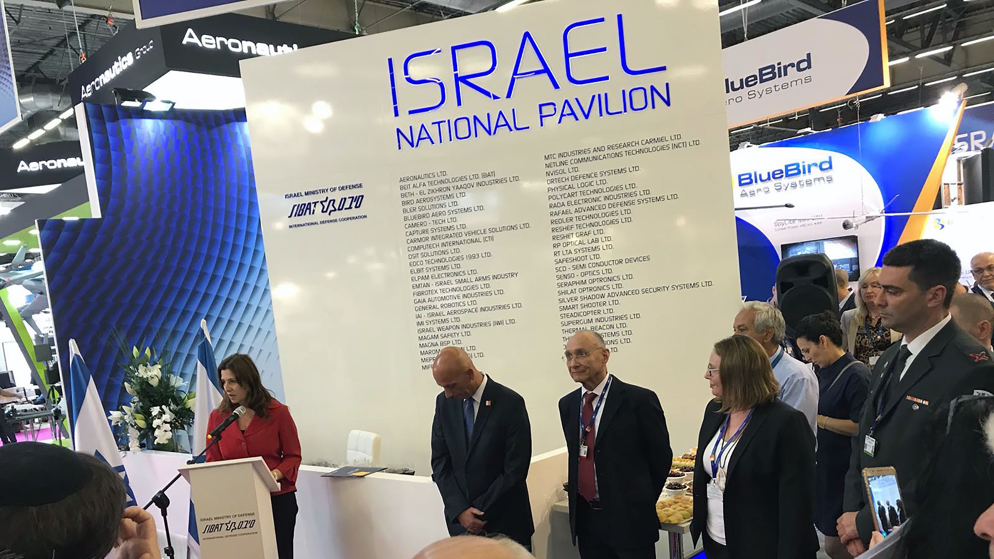 La imagen muestra un evento en un pabellón nacional de Israel. En el centro, hay una mujer hablando en un micrófono, probablemente realizando un discurso. A su derecha, hay dos hombres, uno con un traje oscuro y el otro con una chaqueta más clara. Detrás de ellos, se pueden ver varias personas en la audiencia, enfocadas en el evento. En la pared del pabellón se encuentra una lista que probablemente muestra exhibiciones o empresas asociadas a Israel. El ambiente parece formal y está relacionado con una exposición o congreso.