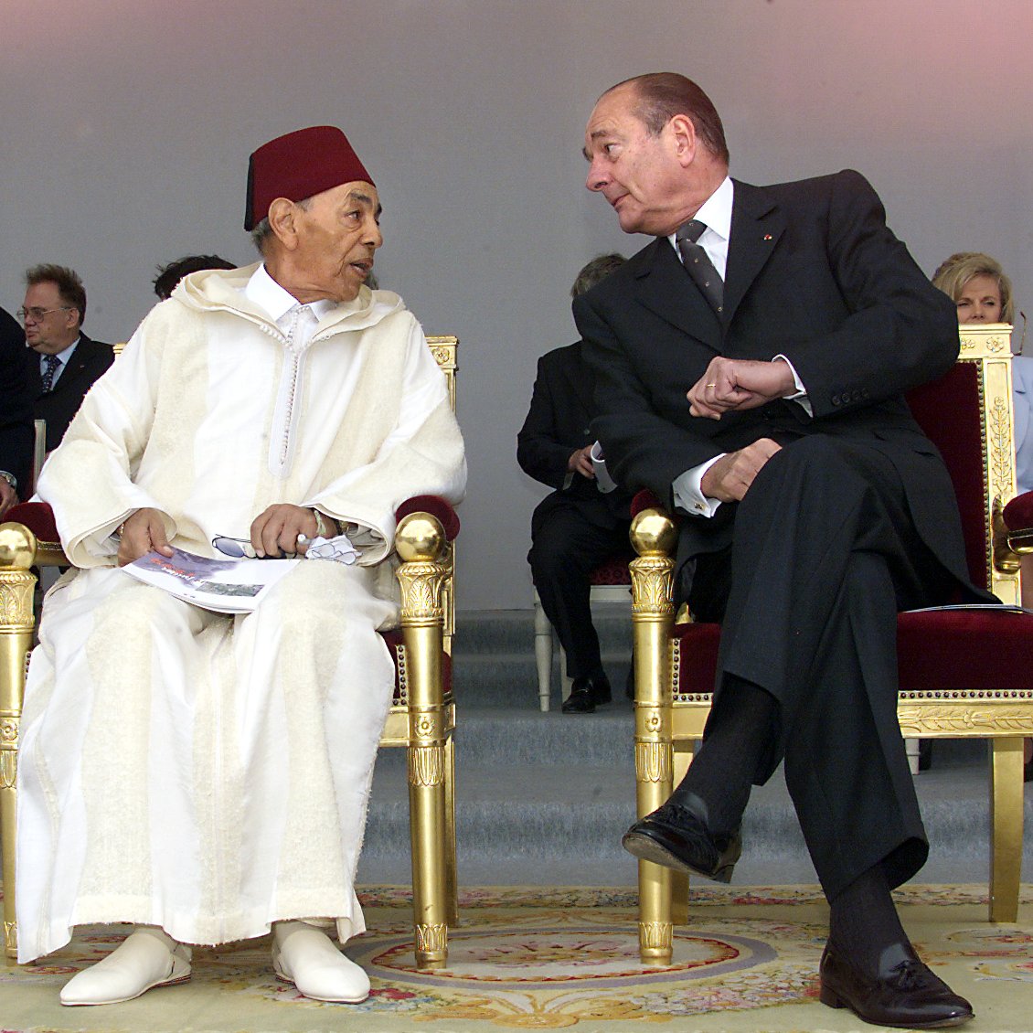 الصورة تظهر شخصين يجلسان على كراسي فاخرة. يبدو أن الحديث يجري بينهما بشكل ودي. الشخص الأول يرتدي زيًا تقليديًا مغربيًا مع طربوش، بينما الشخص الثاني يرتدي بدلة رسمية داكنة. في الخلفية توجد مجموعة من الأشخاص الجالسين، ويبدو أنهم في مناسبة رسمية أو احتفال. الضوء في الصورة ناعم، مما يضيف جوًا من الألفة.