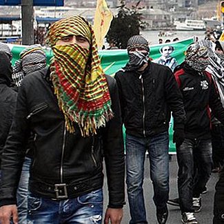 L'image montre une manifestation de foule, où de nombreuses personnes marchent ensemble. Beaucoup d'entre elles portent des masques ou des foulards qui couvrent leur visage, ce qui donne un aspect uniformisé à ce groupe. On constate une ambiance de protestation, probablement liée à une revendication sociale ou politique. En arrière-plan, on aperçoit des drapeaux et des banderoles, indiquant des messages forts. Les participants semblent déterminés, et l'atmosphère semble chargée d'énergie et de tensions.
