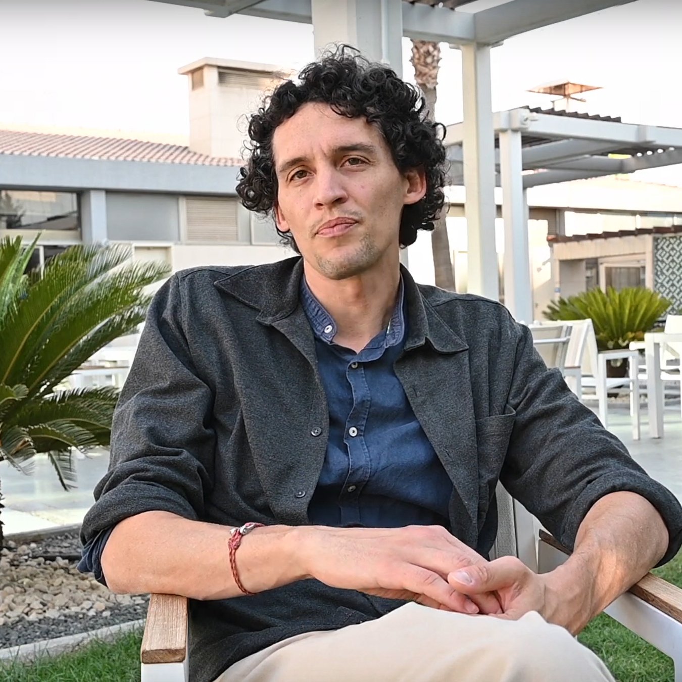 L'image montre un homme assis sur une chaise en bois dans un environnement extérieur. Il a des cheveux bouclés et porte une chemise sombre sur un t-shirt. Il est assis dans un cadre paisible, avec des plantes vertes autour de lui et une terrasse visible en arrière-plan. L'ambiance semble détendue et agréable.
