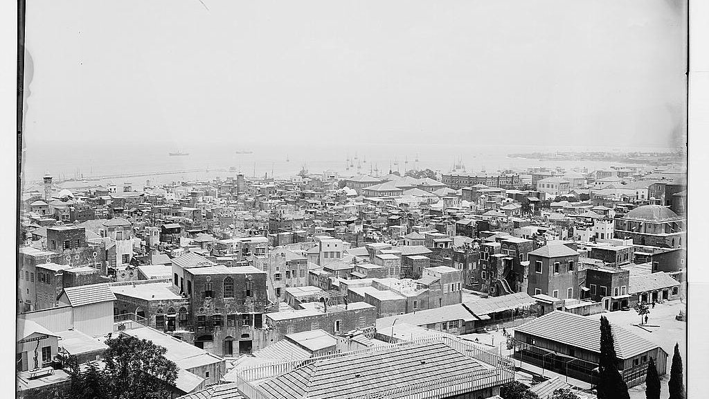 L'image présente une vue panoramique d'une ville côtière. On peut voir de nombreux bâtiments aux toits en tôle, ainsi que des structures en maçonnerie. La mer est visible à l'arrière-plan, avec quelques bateaux amarrés. Les rues semblent étroites et bien dessinées, et il y a des arbres éparpillés dans la zone. L'ensemble donne une impression de densité urbaine et de vie maritime. La photographie semble ancienne, capturant l'architecture et l'ambiance d'une époque révolue.