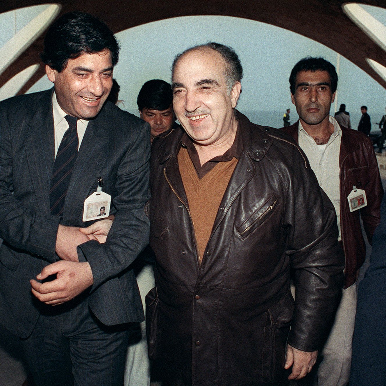 L'image montre un groupe d'hommes évoluant dans un environnement architectural moderne. Deux hommes au premier plan semblent converser de manière amicale, affichant de larges sourires. L'un est vêtu d'une veste en cuir, tandis que l'autre porte une tenue plus formelle. En arrière-plan, d'autres personnes les accompagnent, et tous semblent se déplacer sous une structure aux lignes architecturales caractéristiques. L'atmosphère générale de la photo semble être celle d'une rencontre ou d'un événement significatif.