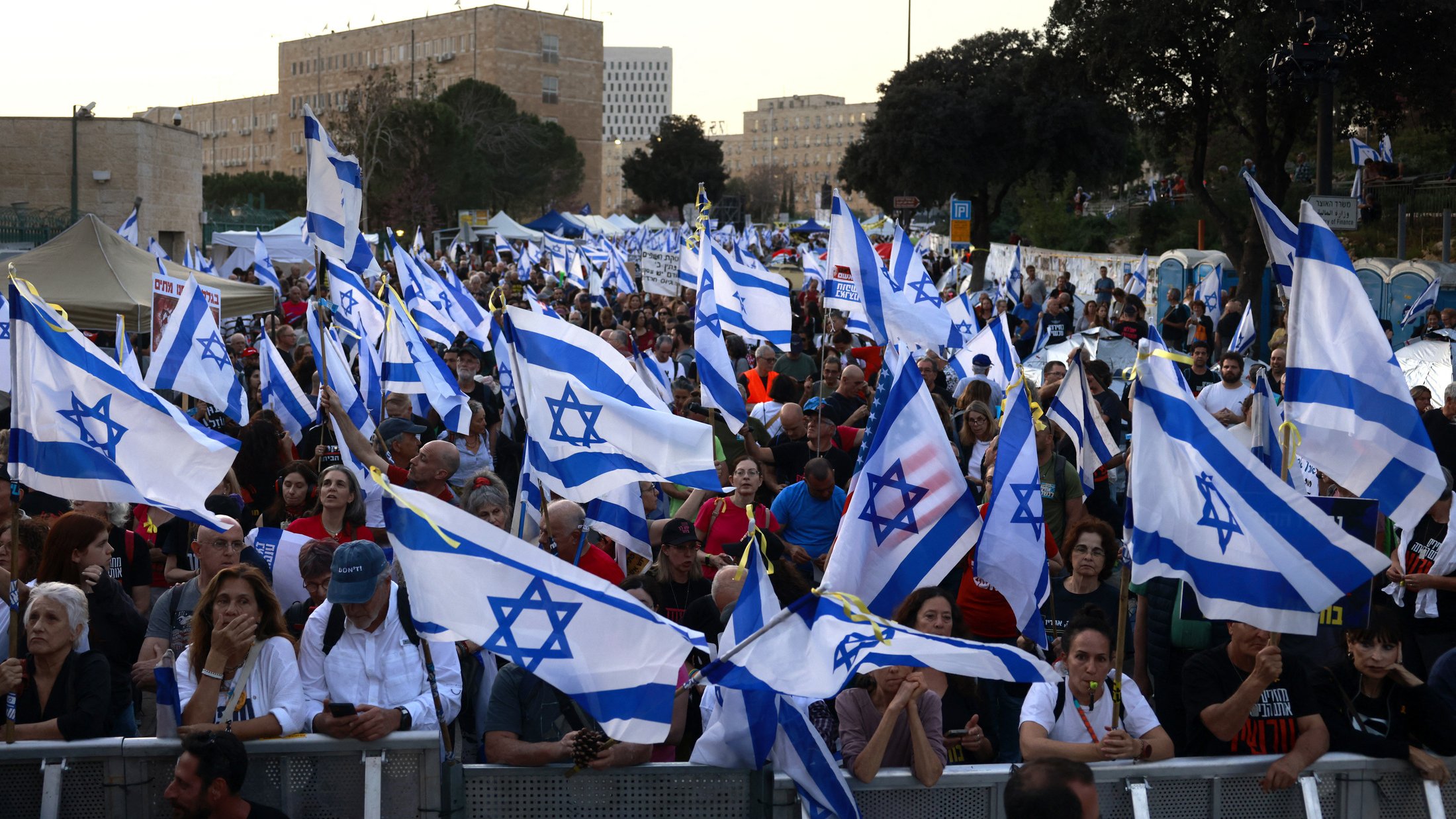 The image shows a large crowd of people gathered together, holding numerous Israeli flags. The atmosphere appears to be part of a demonstration or rally, with many individuals engaged in conversation or focused on the event. The background features buildings and trees, indicating an urban setting. The flags are prominently displayed, creating a sense of unity among the participants.