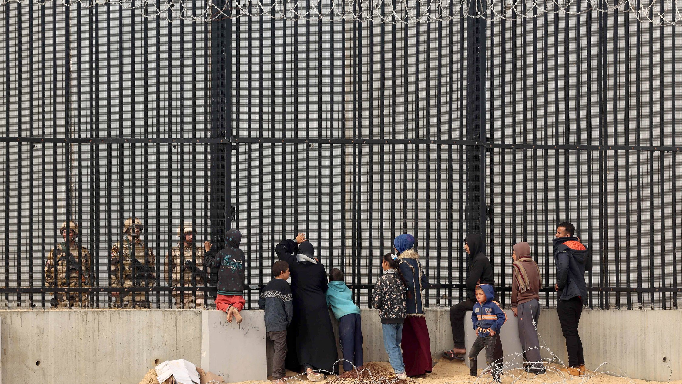 تظهر الصورة مجموعة من الأشخاص الواقفين أمام حواجز حديدية أو سياج مسنن، حيث يواجهون جنود أو عناصر أمنية على الجانب الآخر. يبدو أن هناك أطفالًا ونساء ورجالًا، وبعضهم يحملون أكياسًا أو متعلقات أخرى. البيئة المحيطة موحشة، مع وجود سلك شائك في المقدمة، مما يوضح التوتر والمشاعر القاسية في هذا المشهد.