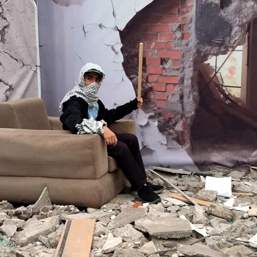 L'image montre une scène de destruction avec des débris éparpillés au sol, tels que des morceaux de béton et des morceaux de verre brisé. Au centre, une personne est assise sur un canapé apparemment plus ou moins intact. Elle porte un foulard autour de la tête et tient un bâton. En arrière-plan, il y a une illustration d'un mur brisé, ajoutant à l'impression de désolation et de chaos de la scène.