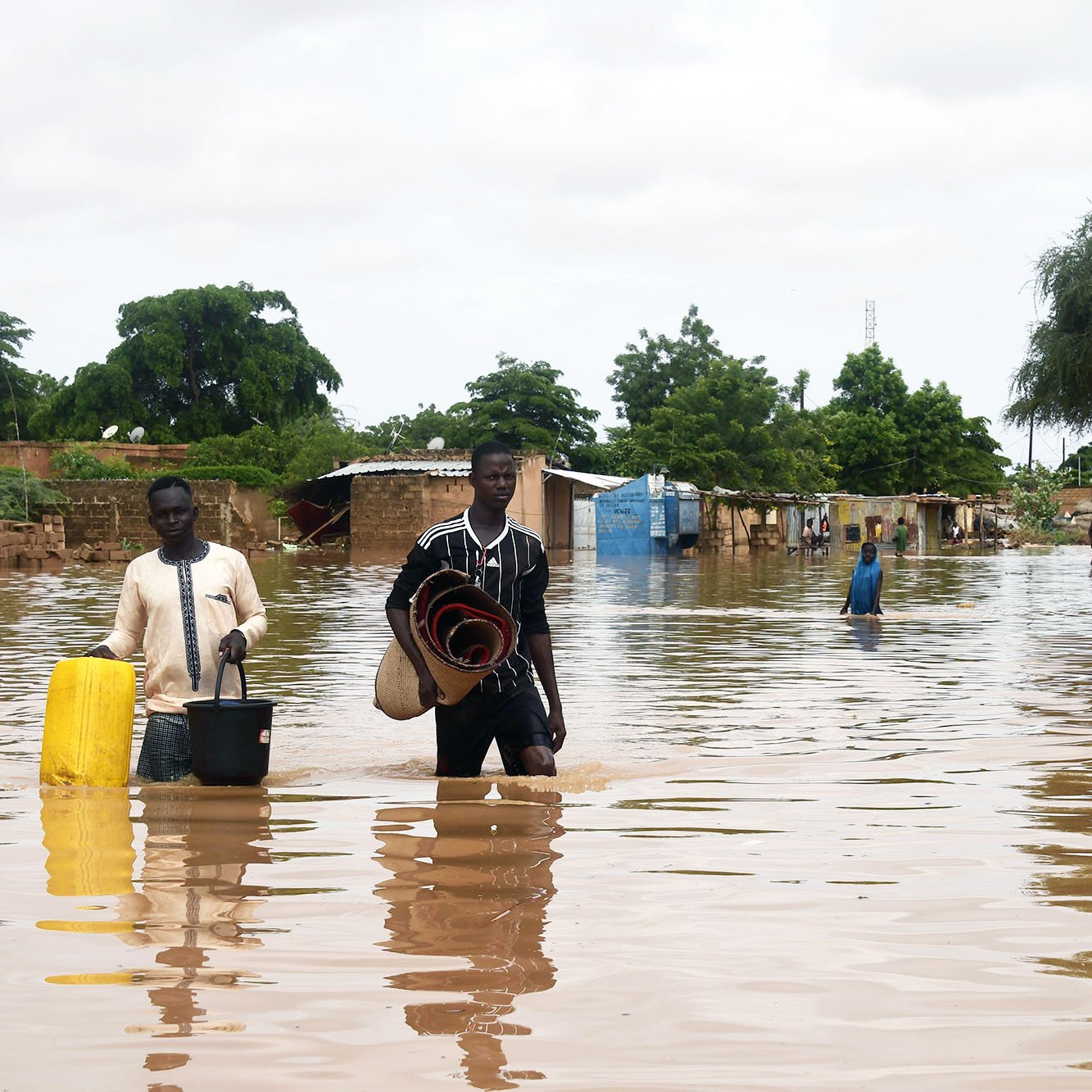 L'image montre une scène de inondation où deux hommes marchent dans une zone inondée. L'eau monte jusqu'à leurs jambes et environ une partie des maisons environnantes est submergée. Les hommes portent des objets, l'un avec un seau et l'autre avec des paniers ou des matelas. En arrière-plan, on peut voir des arbres et des bâtiments partiellement immergés, représentant une situation difficile due aux inondations. Le ciel est nuageux, ce qui suggère des conditions météorologiques instables.