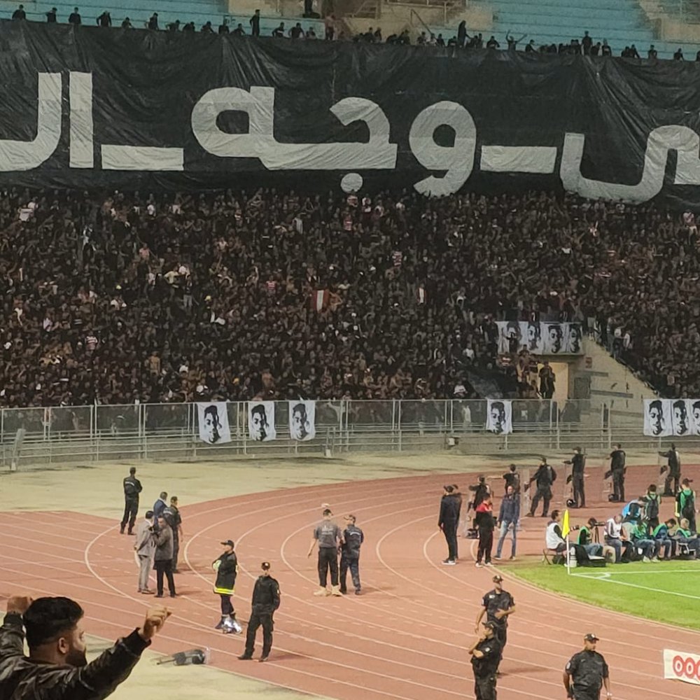 Stade plein de supporters en noir avec une grande banderole en arabe. Énergie et passion.