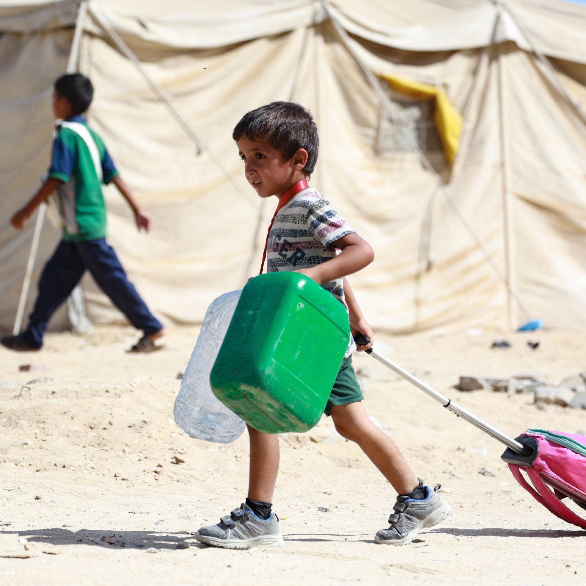 L'image montre un enfant marchant dans un environnement de camp, probablement un camp de réfugiés. Il tire une valise rose avec une poignée, tout en portant un récipient vert dans ses bras. En arrière-plan, on peut voir d'autres enfants et des tentes en toile, symbolisant un cadre de vie précaire. Le sol est poussiéreux et dénudé, ce qui reflète les conditions difficiles de l'endroit. L'enfant a un visage déterminé et semble concentré sur sa tâche.