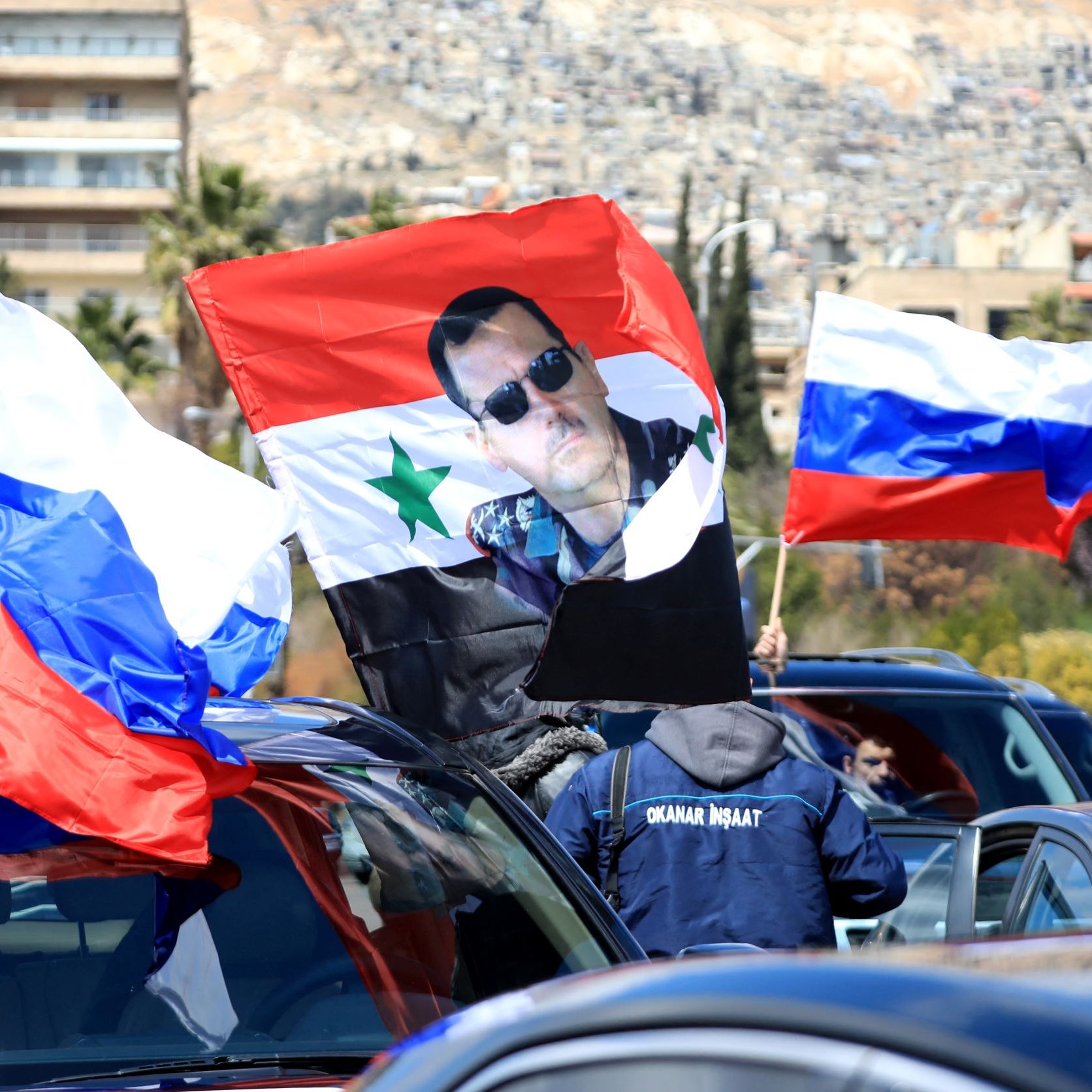 L'image montre une scène dans laquelle des personnes portent des drapeaux russes et un drapeau syrien. Au centre, on peut voir un grand drapeau sur lequel est imprimée une photo d'un homme. La scène semble se dérouler dans une ville, probablement en Syrie, avec des bâtiments en arrière-plan sur une colline. L'atmosphère suggère un rassemblement ou une manifestation, possiblement en soutien à une cause politique.