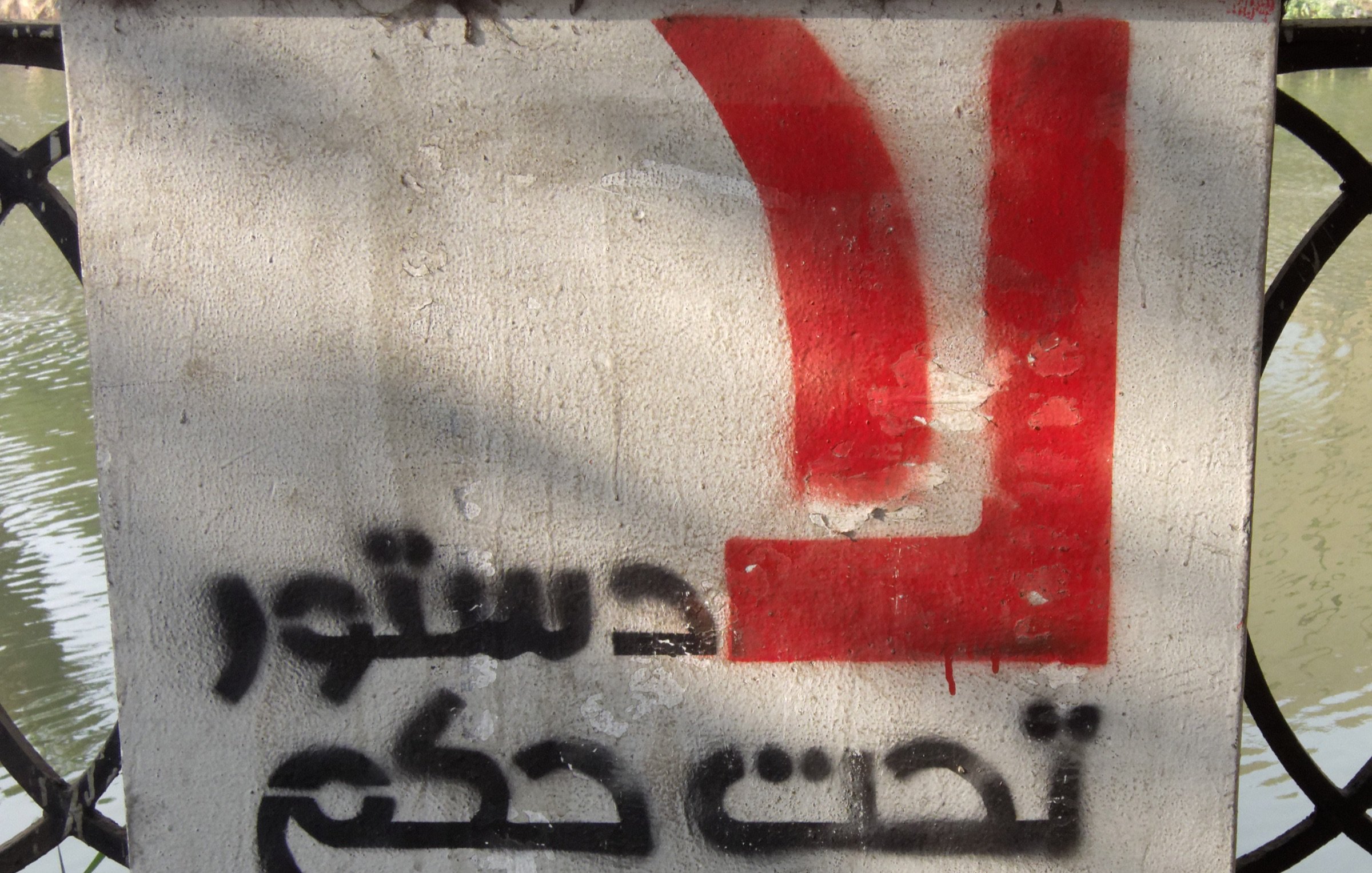L'image montre un panneau sur un mur en béton, avec un fond gris. Sur la partie supérieure, on peut voir un symbole ou une lettre en rouge. En dessous, des mots sont écrits en arabe, et le texte semble faire référence à un sujet politique, évoquant un appel ou une déclaration. Le tout est en contraste avec l'environnement aquatique en arrière-plan.