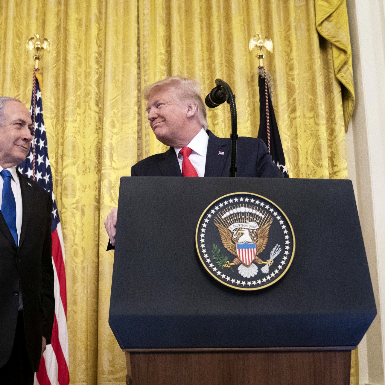 تصویر مربوط به یک مراسم رسمی است که در آن دو نفر در حال گفتگو هستند. در پس‌زمینه، پرده‌های طلایی و پرچم‌های ایالات متحده دیده می‌شوند. یکی از افراد در سمت چپ، لباسی تیره به تن دارد و به شخص دیگر در سمت راست نگاه می‌کند. شخص در سمت راست یک لباس رسمی و یک کراوات به تن دارد و به نظر می‌رسد در حال لبخند زدن است. همچنین بر روی میزی که در مقابل آنها قرار دارد، نشان رسمی ریاست‌جمهوری ایالات متحده وجود دارد. این تصویر به نظر می‌رسد به یک رویداد سیاسی یا دیپلماتیک مربوط باشد.