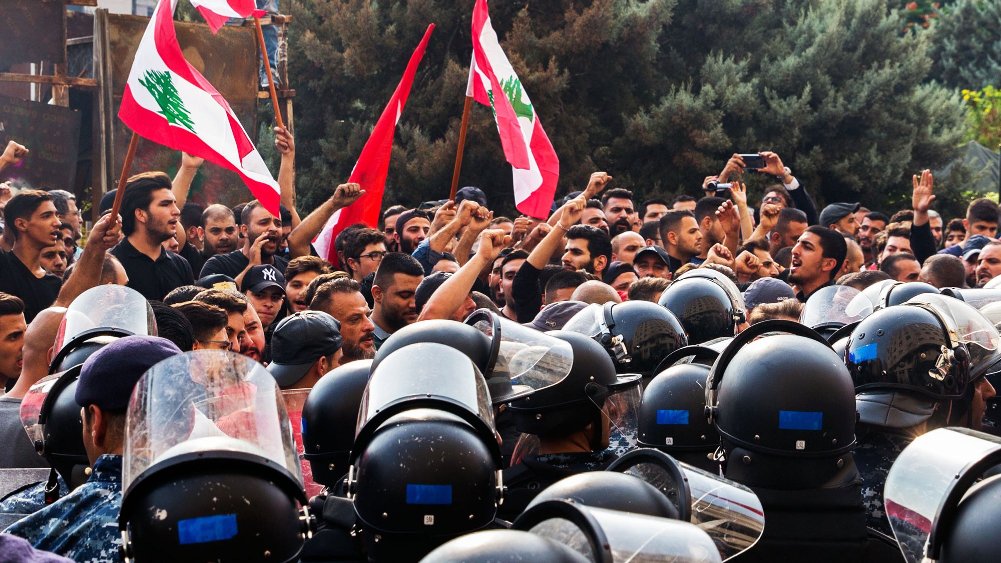 L'image montre une scène de manifestation avec un grand nombre de personnes brandissant des drapeaux libanais. Les manifestants semblent exprimer leurs revendications avec des poings levés, tandis qu'une ligne de policiers en uniforme, équipés de casques et de protections, se tient face à eux. L'ambiance est tense, illustrée par la proximité des manifestants et des forces de l'ordre. Des arbres en arrière-plan complètent le décor de cette confrontation.