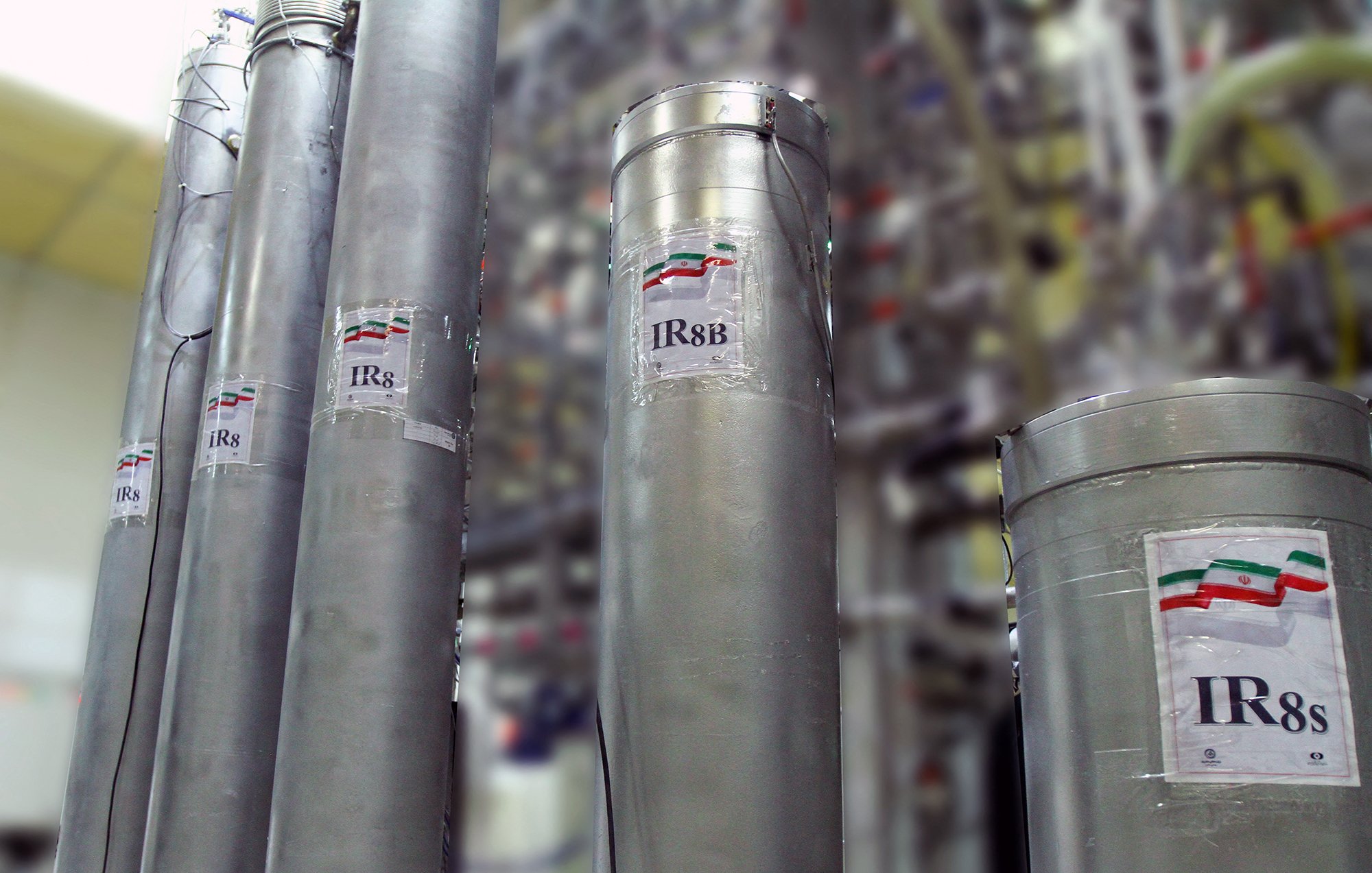 La imagen muestra varios cilindros metálicos dispuestos en un laboratorio o instalación industrial. Cada cilindro tiene una etiqueta que indica "IR8" o "IR8b", probablemente refiriéndose a modelos de centrifugas. En las etiquetas se puede ver una representación de la bandera de Irán. El fondo está desenfocado, pero se pueden observar más estructuras relacionadas con el equipo técnico, sugiriendo un ambiente relacionado con la investigación o la producción.