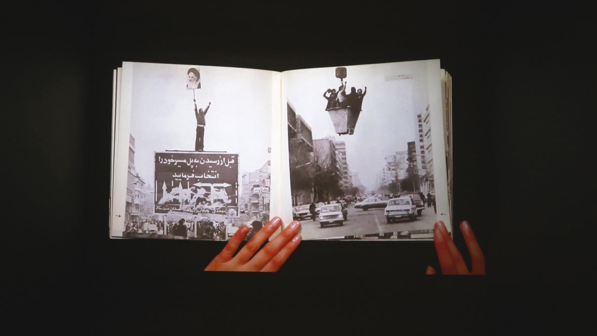 L'image montre un livre ouvert, tenu par des mains, avec des pages affichant des photos en noir et blanc. L'une des pages présente une grande pancarte ou affiche, tandis que l'autre illustre une rue avec des voitures. L'ensemble évoque des thèmes de mouvement, de protestation ou d'événements historiques, en jouant sur le contraste entre les images et le texte visibles. L'arrière-plan est sombre, ce qui met en valeur le livre et les illustrations qu'il contient.