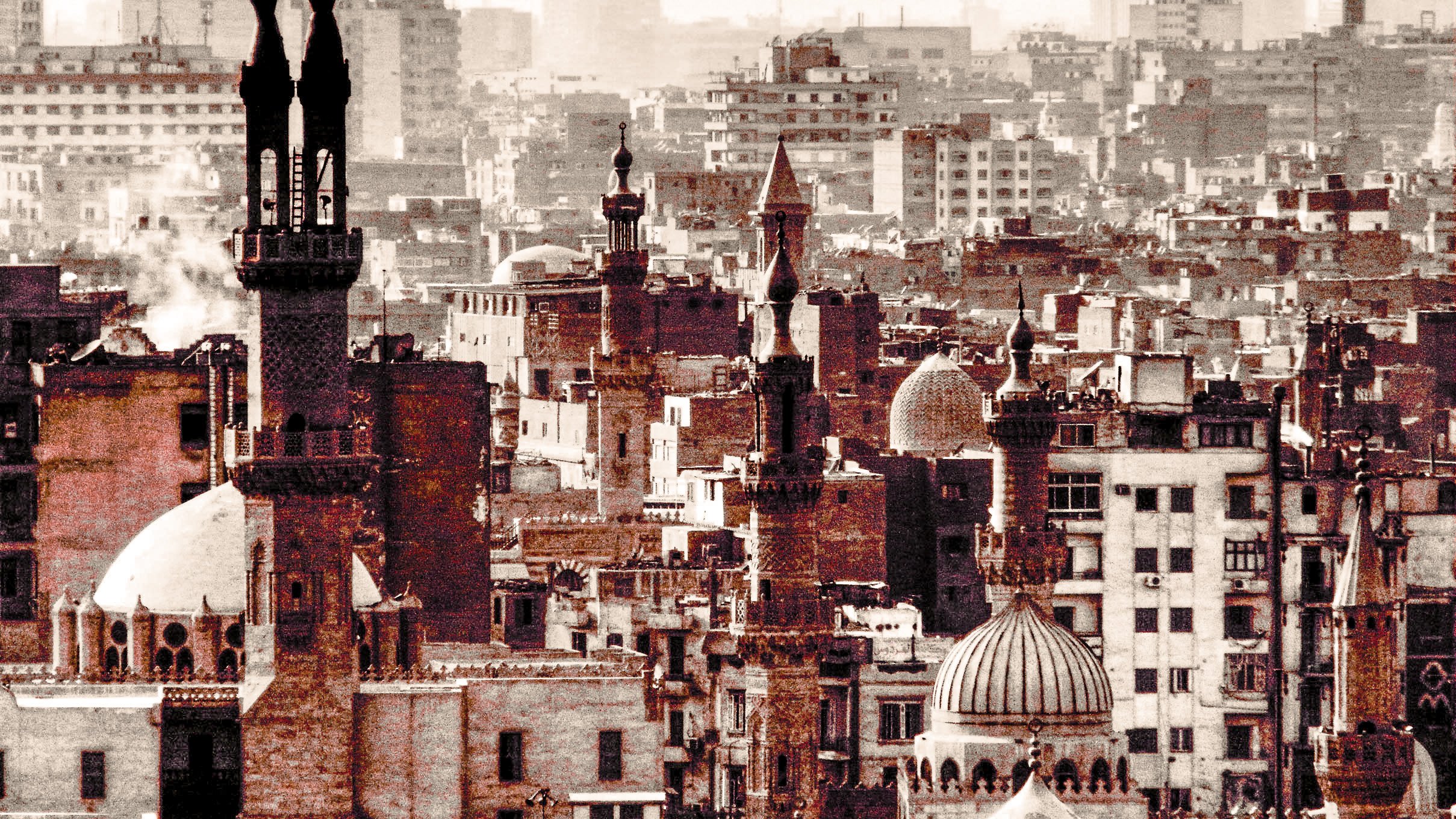 L'image montre un paysage urbain d'une grande ville, probablement au Moyen-Orient. On y distingue plusieurs minarets et dômes de mosquées, reflétant une architecture historique et religieuse. Les bâtiments sont de styles variés, certains ayant des tours élancées. La scène est baignée dans des teintes sèches, ce qui donne une ambiance nostalgique. En arrière-plan, on aperçoit des gratte-ciel modernes, créant un contraste avec l'architecture ancienne au premier plan. Le brouillard donne une impression de profondeur à l'image, soulignant l'intensité de la vie urbaine.