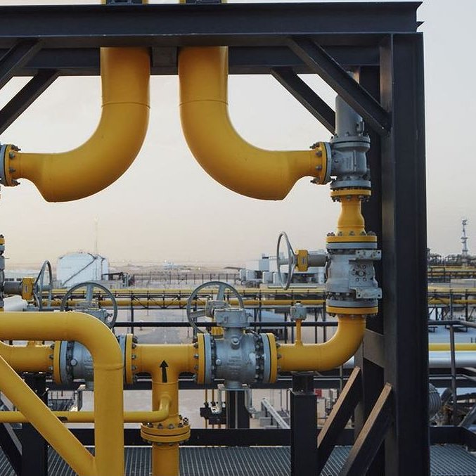 The image features a complex system of industrial piping. Prominent yellow pipes are connected at various angles, showcasing a network of valves and fittings. The background reveals an industrial landscape, with large storage tanks and equipment visible. The overall scene conveys a sense of heavy machinery and infrastructure typically found in oil or gas facilities. The sky appears clear, indicating it might be a time of day either in the morning or late afternoon.
