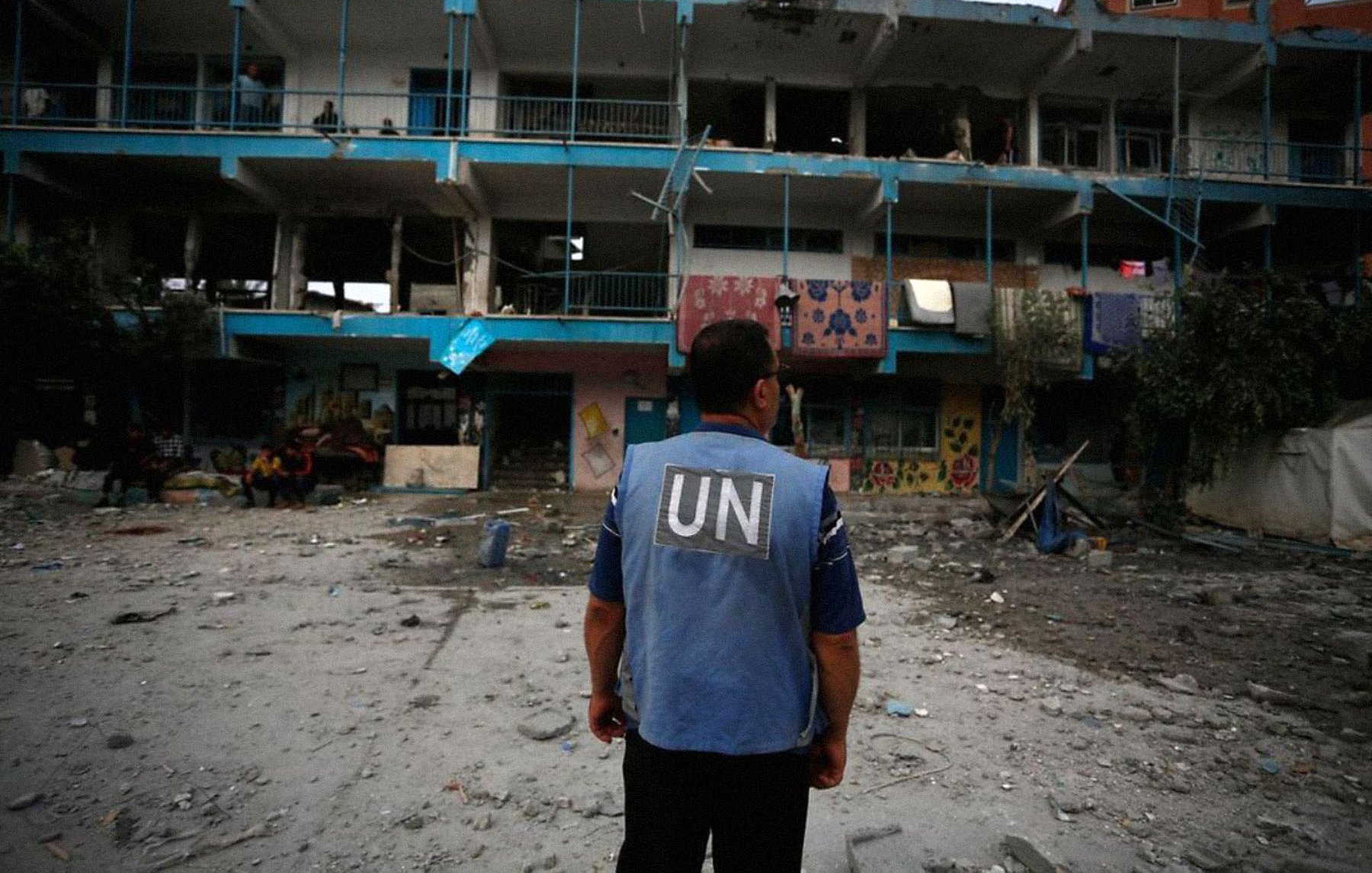 L'image montre un homme portant un gilet de l'ONU, observant un bâtiment endommagé. Le bâtiment a des murs peints et des balcons, mais il est en ruine, avec des fenêtres brisées et des débris éparpillés autour. L'atmosphère est sombre, suggérant une situation de conflit ou de crise, avec d'autres personnes visibles en arrière-plan dans un environnement chaotique.