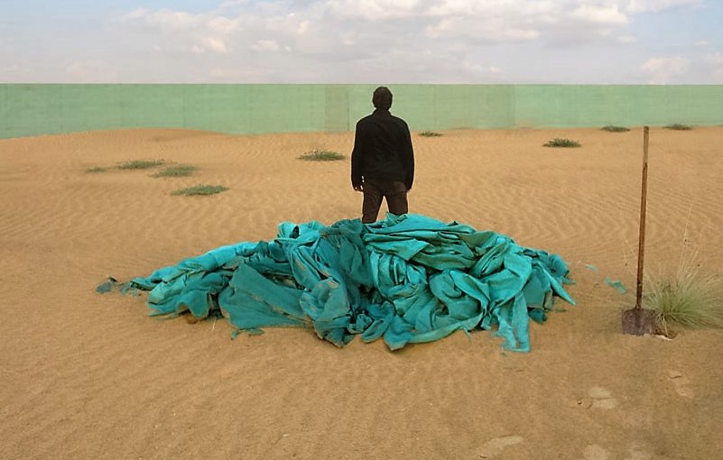 L'image montre un paysage désertique avec des dunes de sable sous un ciel partiellement nuageux. Au premier plan, un homme se tient dos à la caméra sur un monticule de tissus turquoise. En arrière-plan, on aperçoit un mur vert qui contraste avec le sable. L'ensemble de la scène évoque une atmosphère d'isolement et de contemplation.