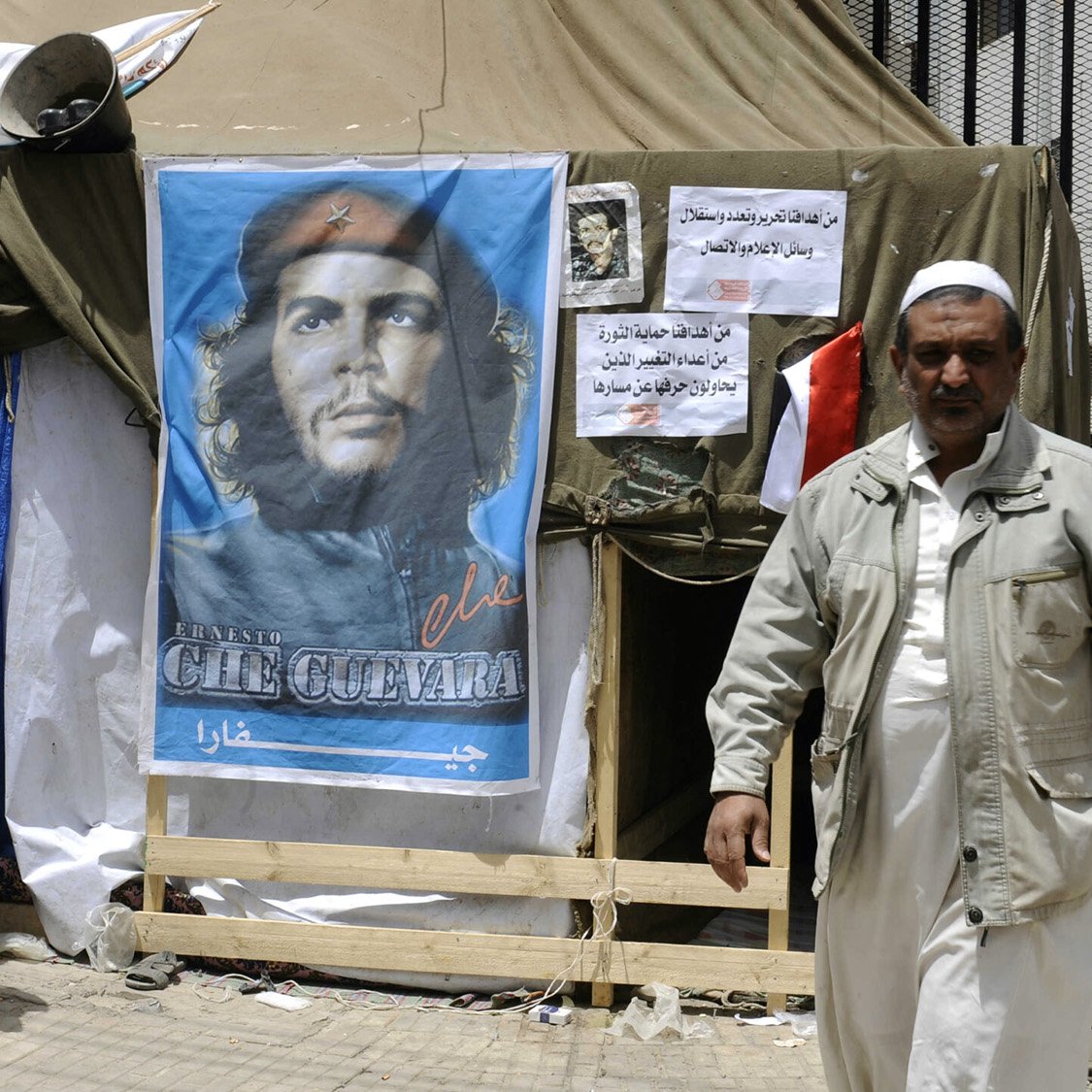 L'image montre un campement avec une tente, devant laquelle se trouve une affiche représentant un homme avec un béret, souvent associé à Che Guevara. À côté de l'affiche, on peut voir des affiches supplémentaires et une personne marchant, vêtue d'une tenue clair. L'ambiance semble refléter un contexte politique ou social, probablement avec des éléments de protestation ou de revendication.