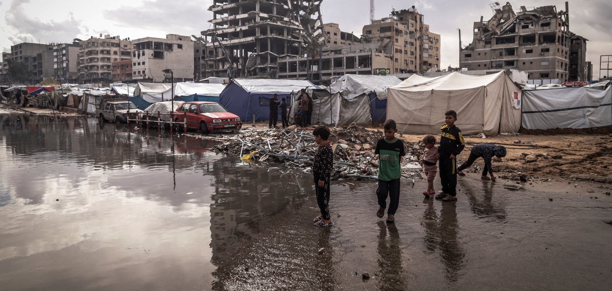 Des enfants jouent près de tentes dans un camp, avec un bâtiment en ruines en arrière-plan.