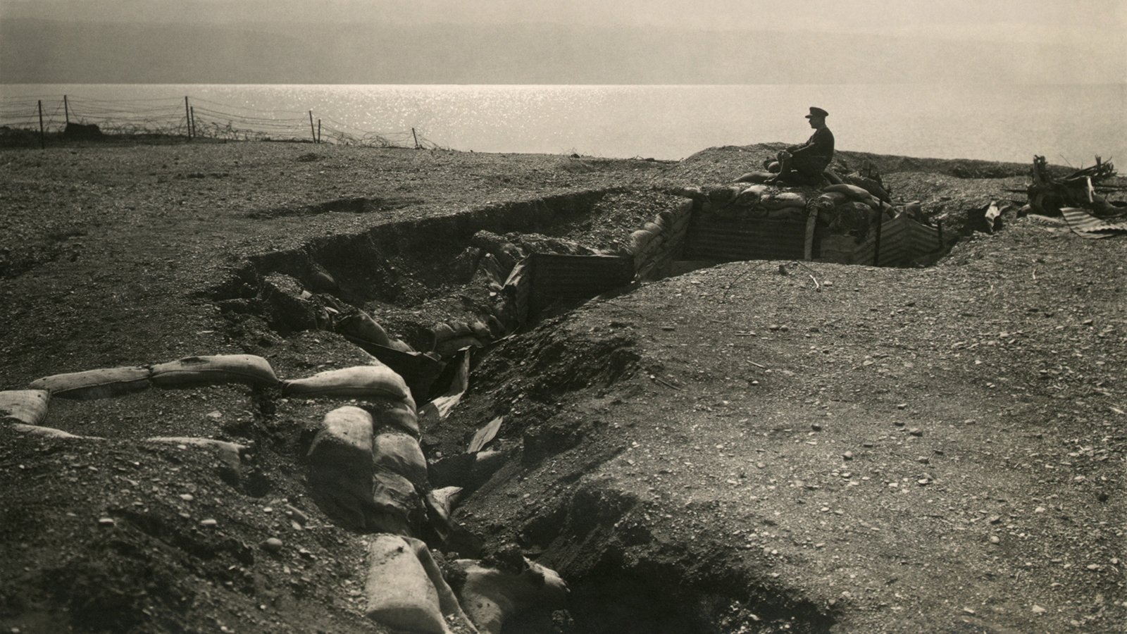 L'image montre une tranchée militaire dans un paysage aride. On peut voir une personne assise sur le sommet d'un rempart en sable, observant les environs. La tranchée est entourée de sacs de sable, et l'horizon est marqué par un plan d'eau au loin. L'atmosphère est sombre et pourrait évoquer une période de conflit, comme la Première ou la Seconde Guerre mondiale. L'éclairage donne à la scène un aspect nostalgique et témoin de l'histoire.