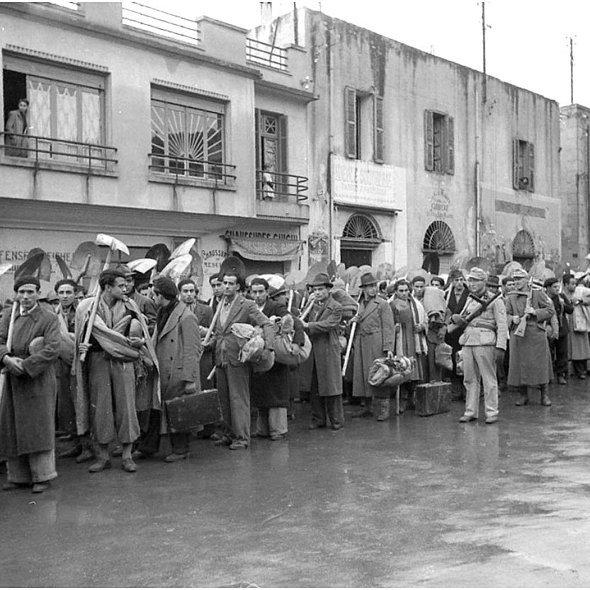 الصورة تظهر مجموعة من الأشخاص في صف طويل، يبدو أنهم ينتظرون شيئًا ما. يرتدون ملابس شتوية، ويحملون حقائب وأشياء أخرى. في الخلفية، يمكن رؤية مبانٍ تاريخية بشرفات ونوافذ. الجو يبدو هادئًا، ولكن المحيط يوحي بوجود حدث مهم أو تجمع. قد تعكس الصورة فترة زمنية معينة أو حدث اجتماعي.