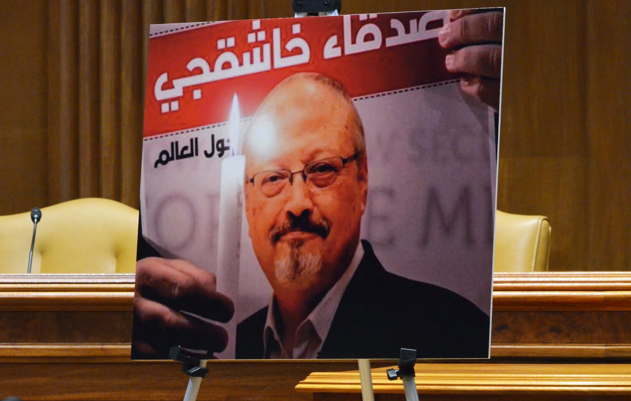 The image features a large poster of a man holding a candle, set against a background with text in Arabic. The poster is likely used in a context related to remembrance or tribute. The setting appears formal, possibly in a legislative or governmental chamber, indicated by the presence of chairs and the overall decor. The image emphasizes themes of justice and memory.