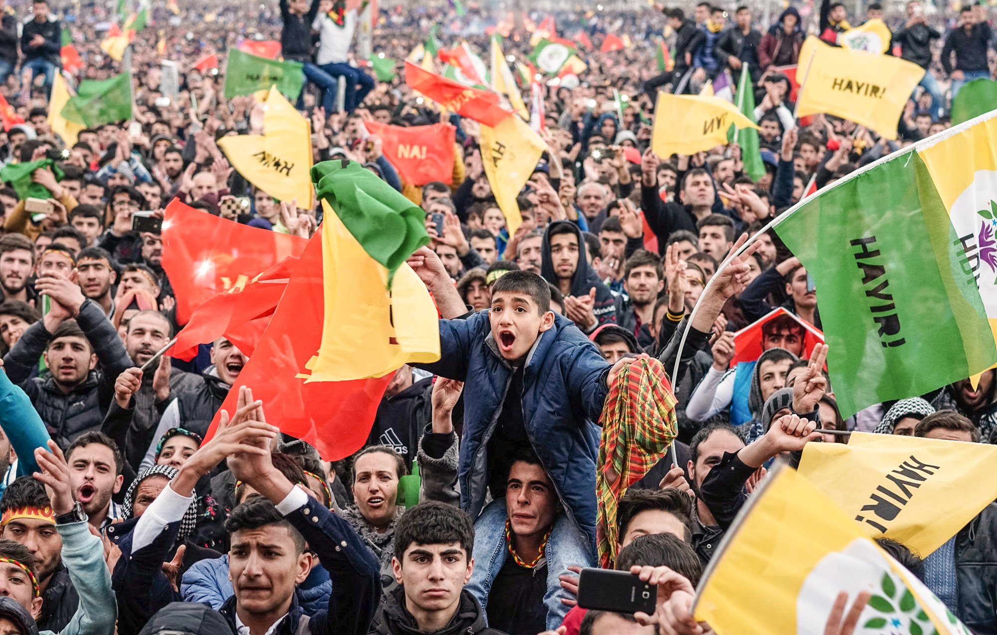 L'image montre une grande foule rassemblée, manifestant avec enthousiasme. Les gens lèvent des drapeaux colorés, dont certains semblent être aux couleurs rouge, jaune et vert. On peut voir un jeune garçon au premier plan exprimant sa joie et son excitation. L'atmosphère est festive, et les participants semblent engagés dans une cause commune, remplis d'énergie et de détermination.