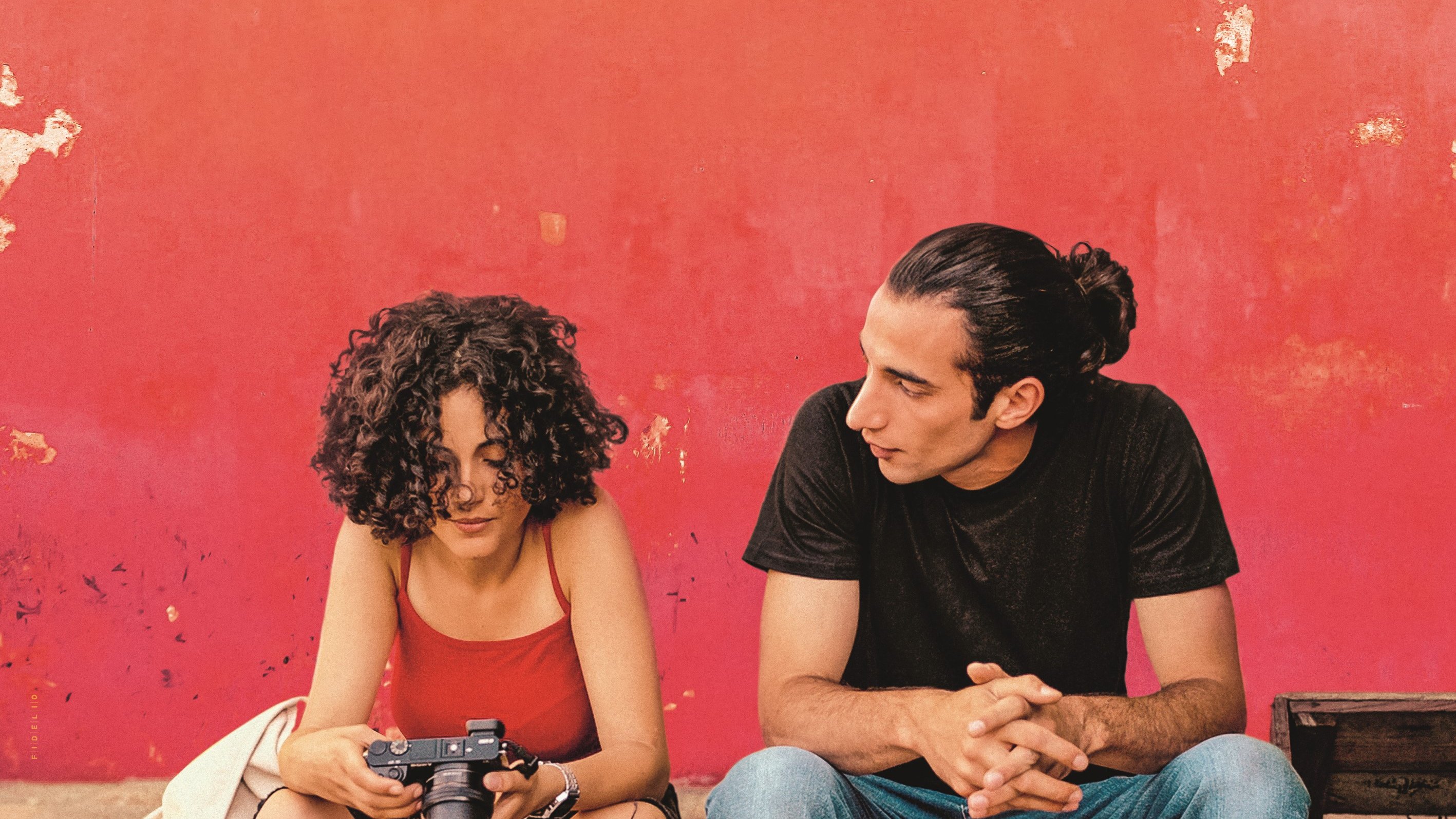 L'image montre deux personnes assises devant un mur rouge écaillé. La première personne, une femme aux cheveux bouclés, tient un appareil photo et semble concentrée sur son écran. Elle porte un débardeur rouge et un gilet clair. À côté d'elle, un homme avec des cheveux longs attachés en chignon la regarde attentivement. Il est vêtu d'un t-shirt noir et de jeans. L'atmosphère paraît détendue et intime, comme s'ils étaient en pleine conversation ou en train de partager un moment.