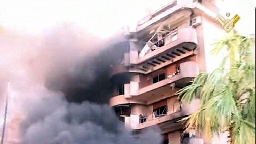 L'image montre un bâtiment en proie à un incendie. De la fumée noire s'échappe des fenêtres, tandis que l'extérieur du bâtiment semble endommagé. Des éléments de végétation, comme un palmier, sont visibles au premier plan, contrastant avec la scène chaotique et la destruction. Le ton général de l'image est sombre, mettant en évidence l'urgence et la gravité de la situation.