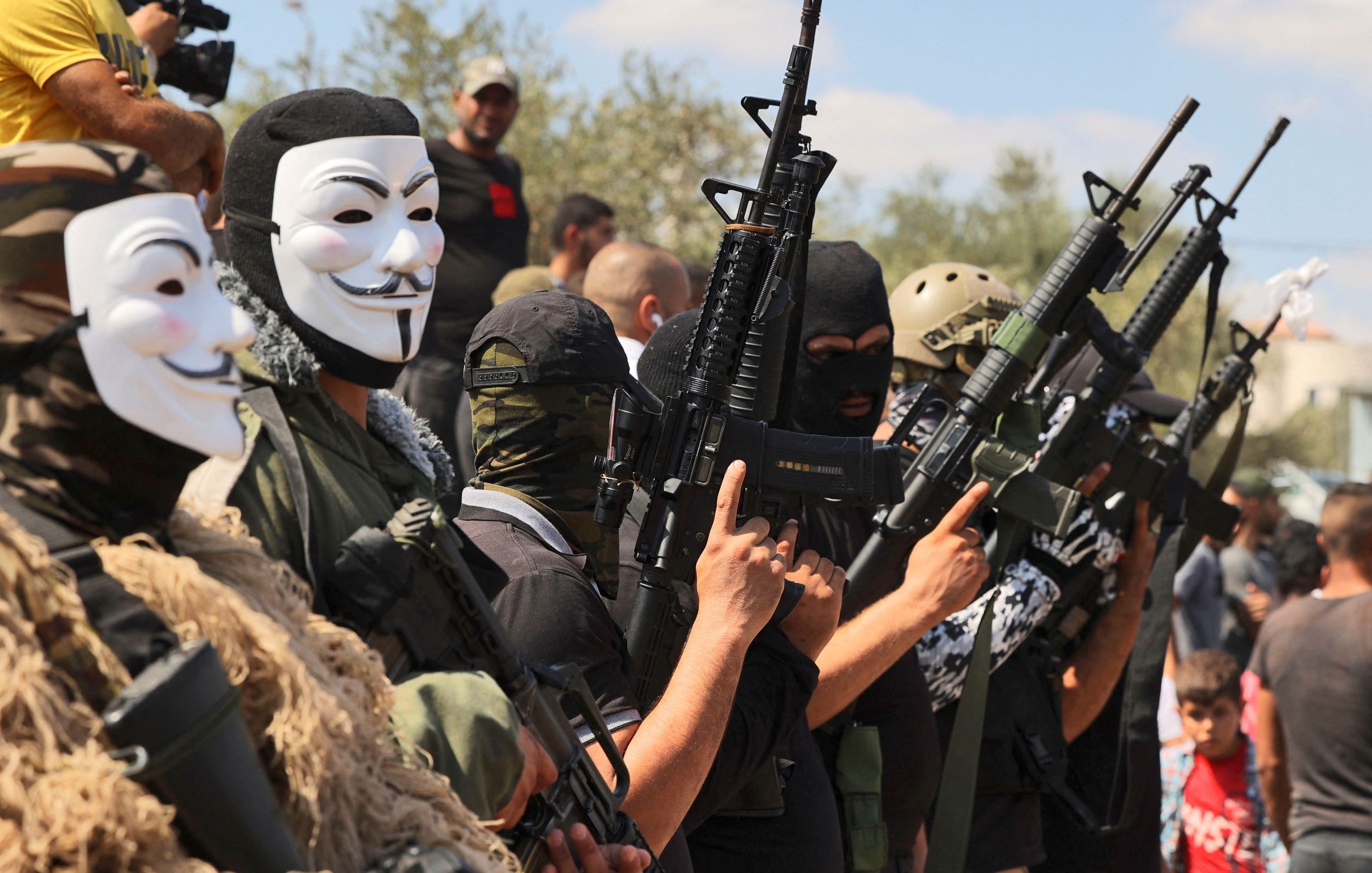 L'image montre un groupe de personnes portant des masques et des vêtements militaires, tenant des fusils. Les visages des individus sont principalement cachés, et ils semblent formés en ligne, chacun montrant une arme. En arrière-plan, on aperçoit d'autres personnes observant la scène. L'atmosphère semble tendue et engagée.