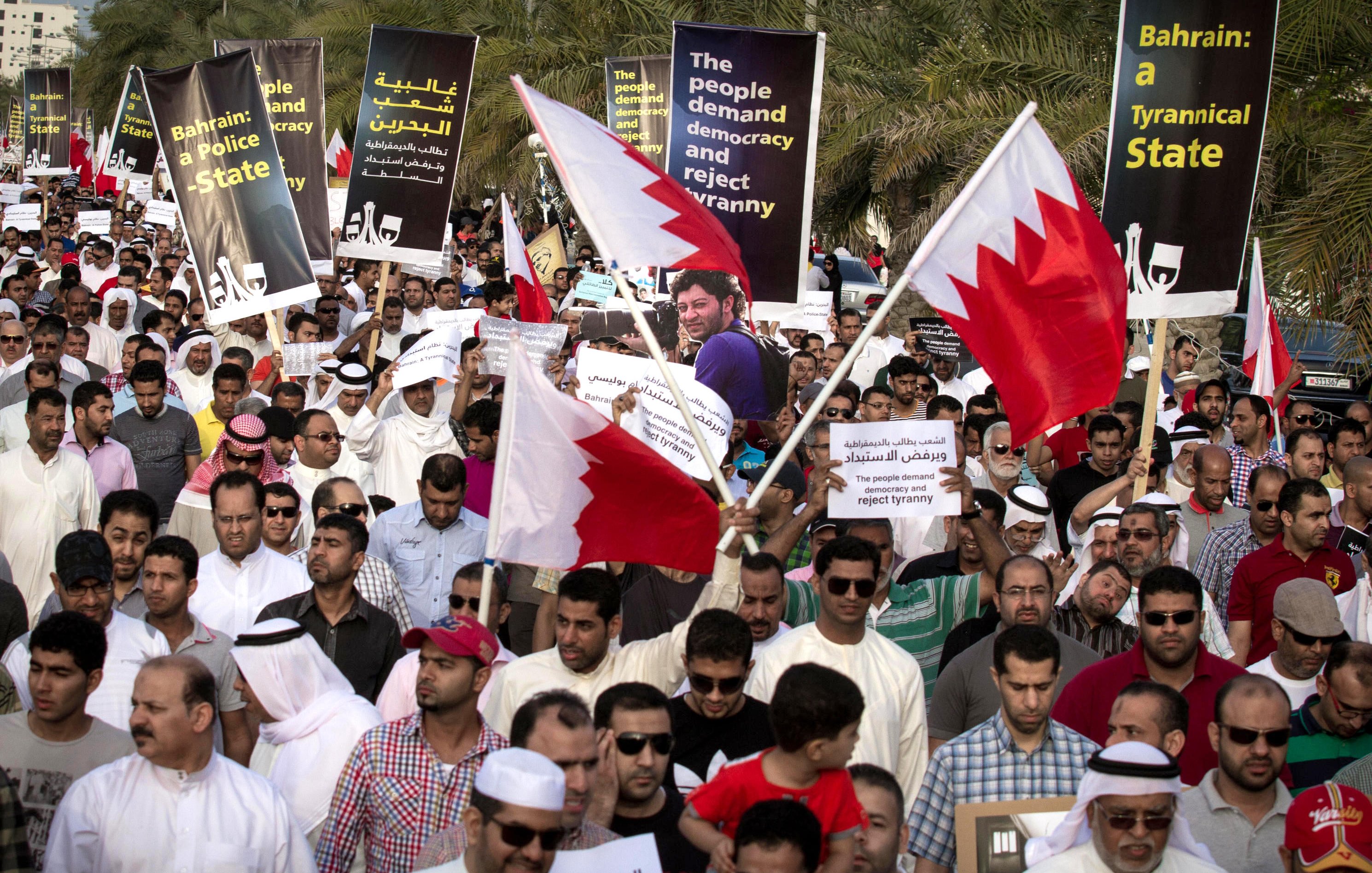 L'image montre une grande foule de manifestants rassemblés dans une rue, tenant des pancartes et des drapeaux de Bahreïn. Les manifestants semblent exprimer des revendications politiques, demandant la démocratie et critiquant le régime en place. On peut voir des gens de différentes origines et genres, tous unis dans leur volonté de changement, avec des expressions déterminées sur leurs visages. L'environnement témoigne d'un climat de mobilisation et de protestation collective.