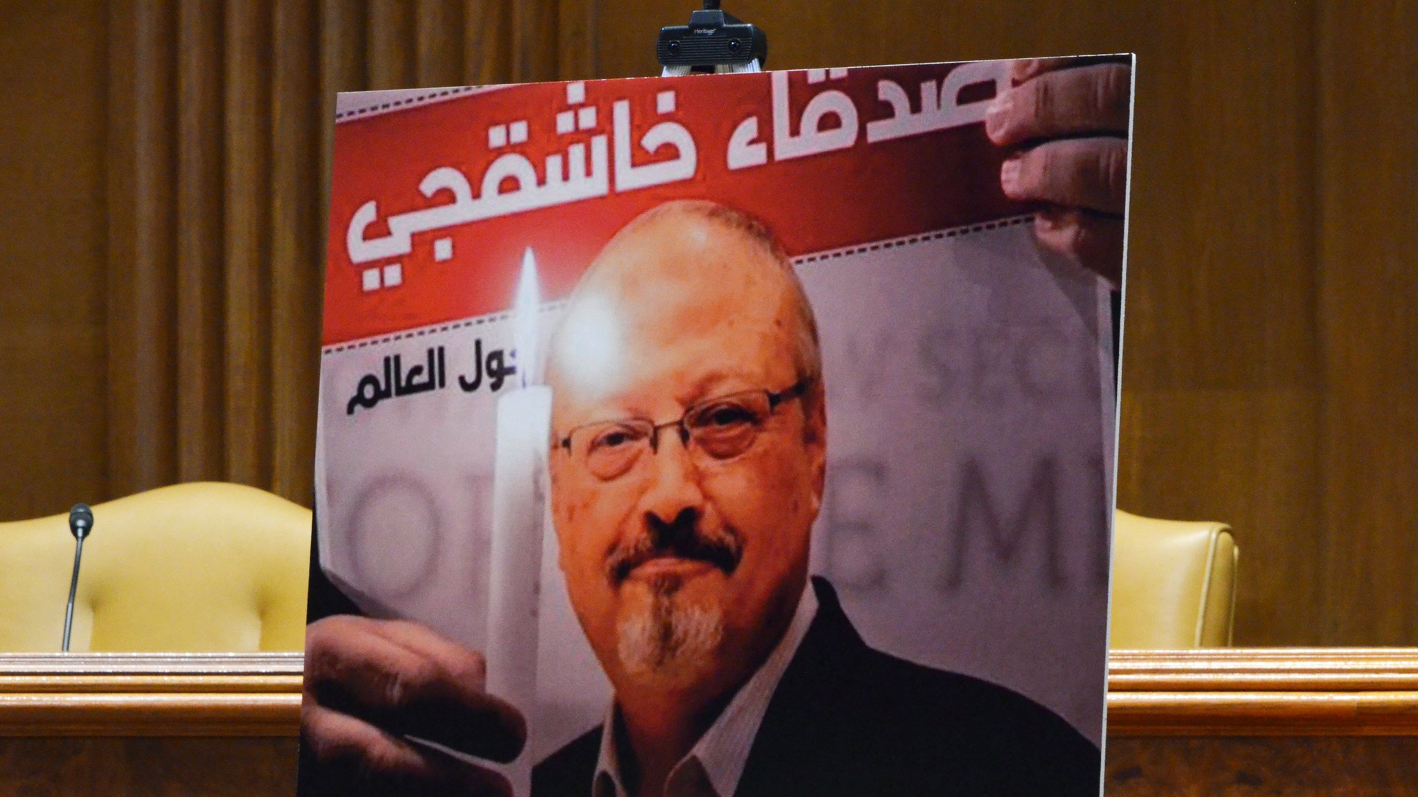 الصورة تظهر لوحة تحمل صورة لصحفي وهو يحمل شمعة مضاءة. خلفه يوجد نص عربي يرد فيه اسم "جمال خاشقجي". يبدو أن اللوحة تم استخدامها في مناسبة لتسليط الضوء على قضيته أو إحياء ذكراه، حيث يعتلي الصورة تعبير عن التضامن أو الاحتجاج. في الخلفية، هناك إعداد رسمي مع مقاعد فارغة.