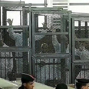 L'image montre un tribunal avec des accusés placés derrière des cages en métal ou des enclos. On aperçoit des personnes à l'intérieur de ces enclos, certaines levant la main. Il y a également des membres des forces de l'ordre en uniforme, observant la scène. L'environnement semble austère, et la lumière naturelle pénètre par des fenêtres en hauteur.