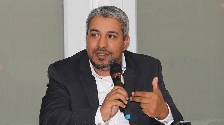 L'image montre un homme assis à une table, tenant un microphone. Il semble engager une discussion ou faire une présentation. Il porte un costume sombre avec une chemise claire et a des cheveux grisonnants. L'environnement autour de lui semble être celui d'une conférence ou d'un événement formel, avec une lumière claire et des fenêtres en arrière-plan.